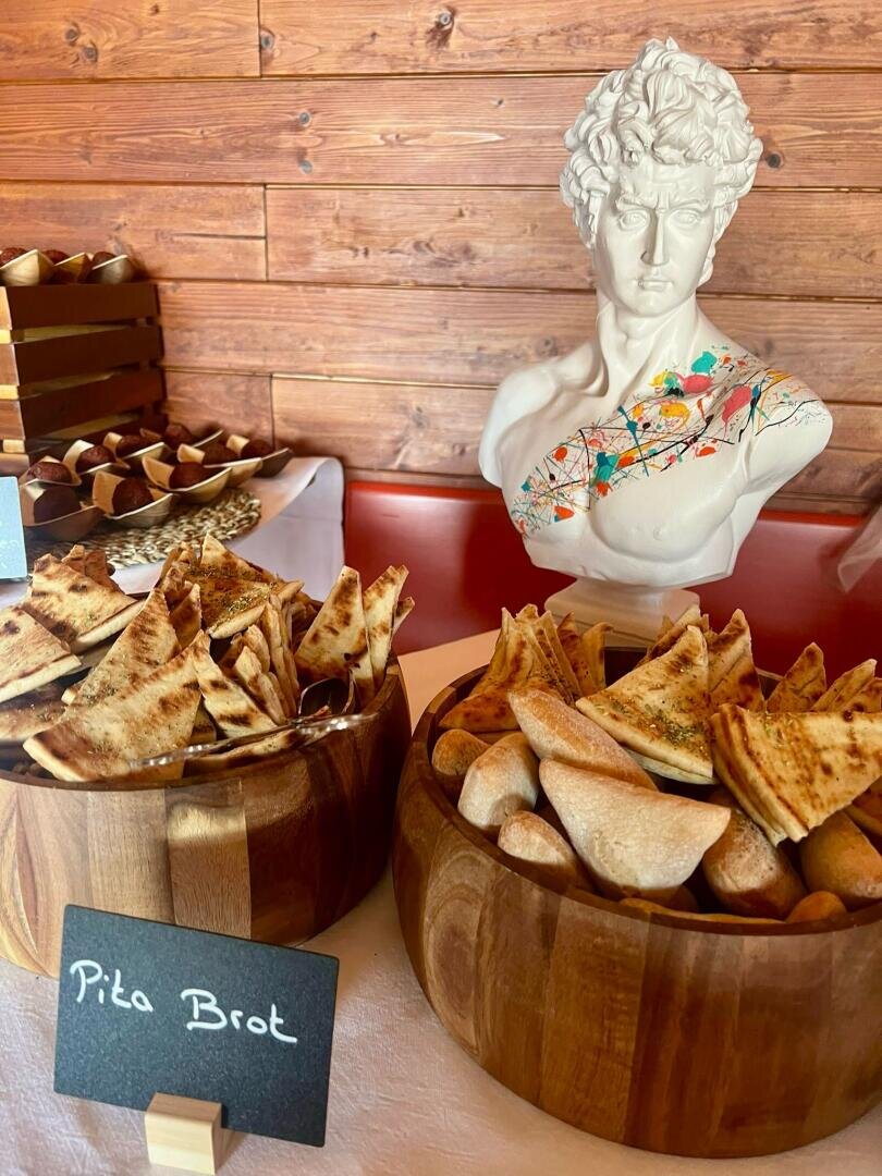 Auf einem Tisch stehen zwei mit Fladenbrotstücken gefüllte Holzschalen neben einem Kreideschild mit der Aufschrift "Pita Brot". Im Hintergrund steht eine weiße Büstenstatue mit bunten Farbspritzern auf der Brust.