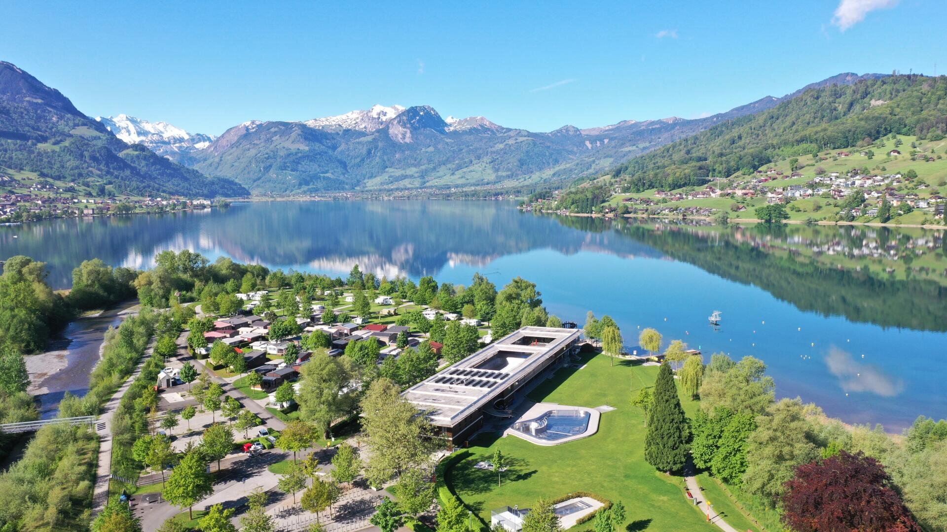 Luftaufnahme eines Seeufers mit einem modernen Gebäude, grünen Bäumen, einem Campingplatz und Bergen im Hintergrund. Der klare blaue See spiegelt die umliegende Landschaft wider, und die Häuser des Dorfes sind auf den Hügeln verteilt.
