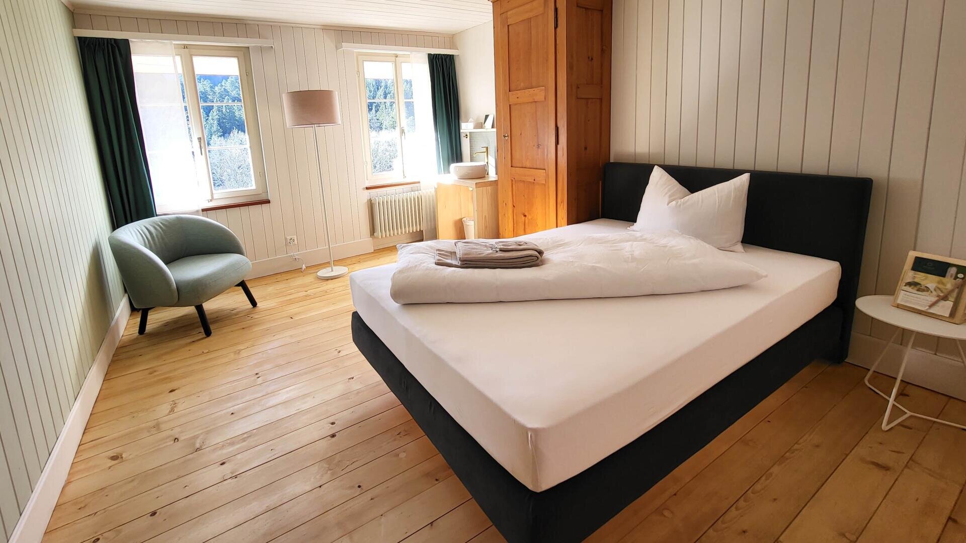 Helles, minimalistisches Schlafzimmer mit einem Doppelbett, weißer Bettwäsche, gefalteten Handtüchern, Holzboden, grünem Sessel, Stehlampe, Holzschrank, kleinem weißen Beistelltisch und zwei Fenstern mit grünen Vorhängen und Blick nach draußen.