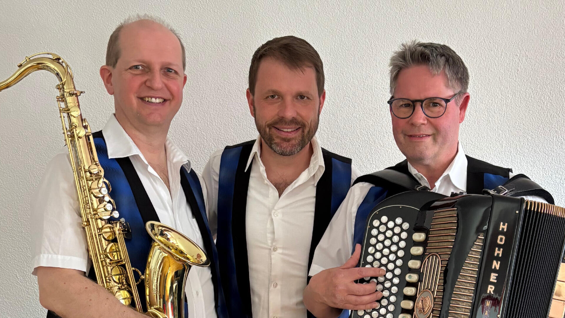 Drei Männer in weißen Hemden und blauen Westen stehen zusammen. Einer hält ein Saxophon, ein anderer ein Akkordeon, und der dritte steht lächelnd in der Mitte. Sie scheinen Musiker zu sein, die für ein Gruppenfoto posieren.