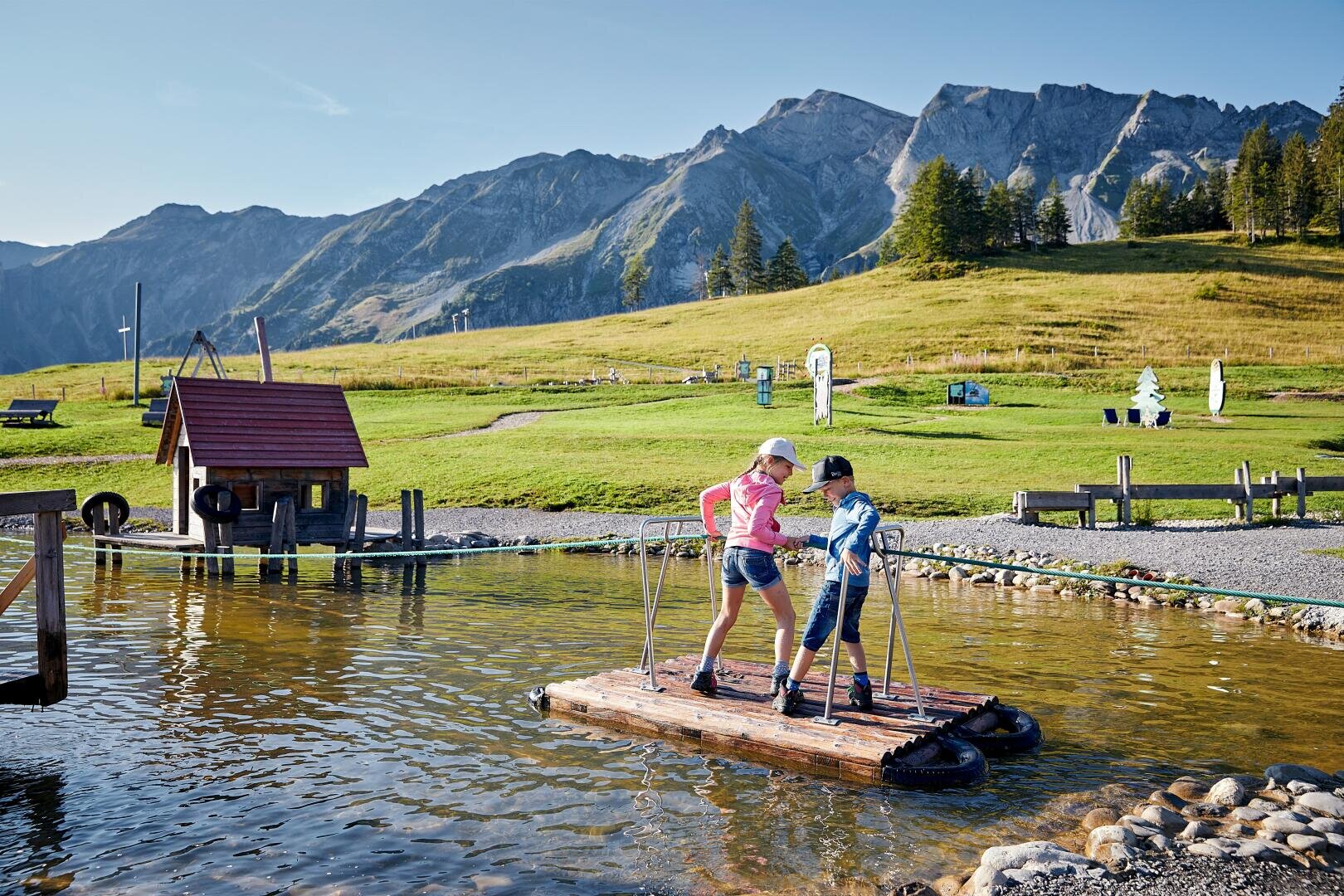Zwei Kinder stehen auf einem Holzfloß in einem kleinen Teich und ziehen sich mit Seilen über das Wasser. Hinter ihnen sind grasbewachsene Felder, eine kleine Hütte und hohe, felsige Berge unter einem blauen Himmel zu sehen.