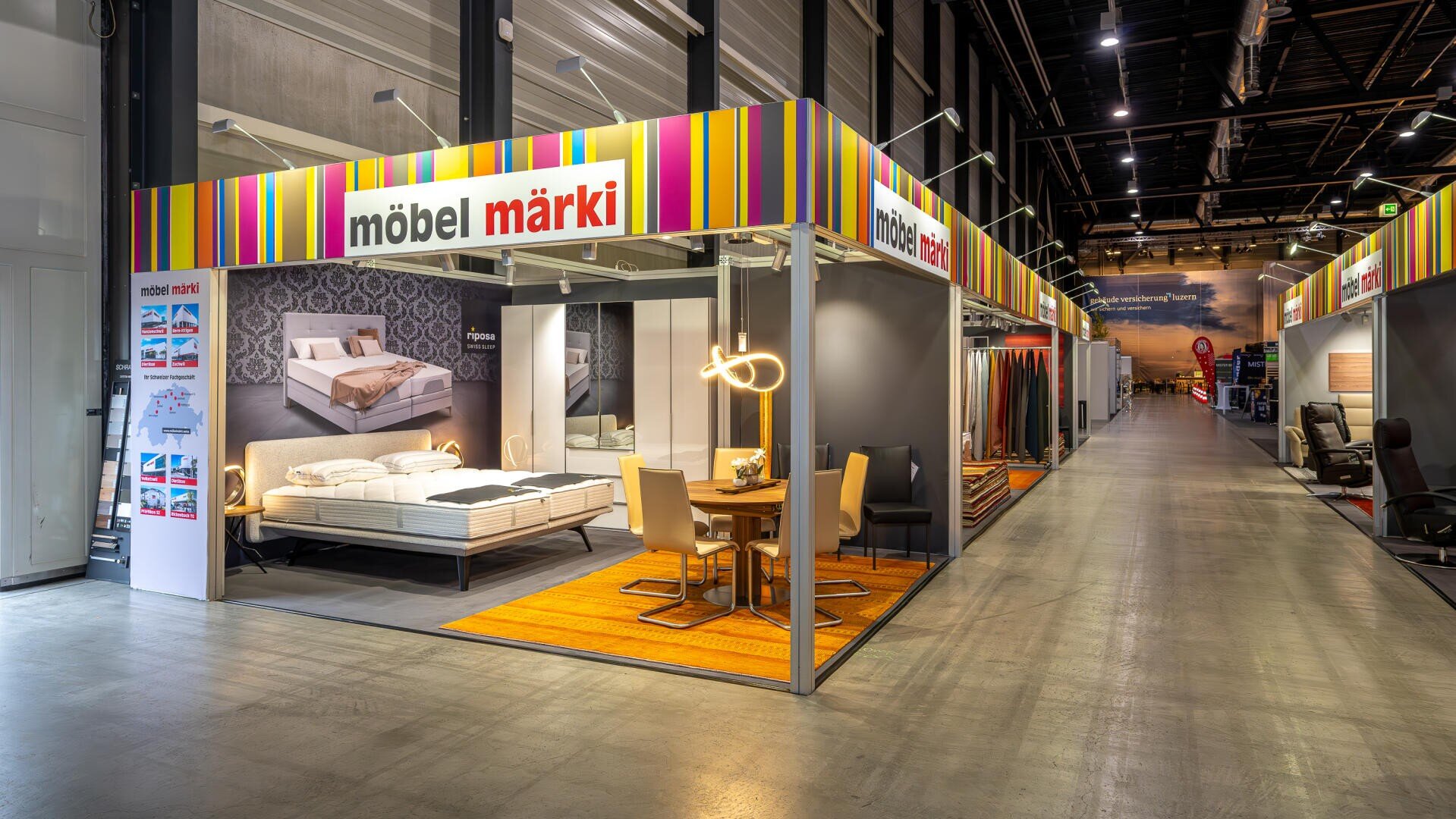 In einem hell erleuchteten Möbelausstellungsraum stehen Betten, ein Esstisch, Stühle und moderne Leuchten unter einem bunt gestreiften möbel märki-Schild, weitere Ausstellungsstände säumen einen breiten Korridor.
