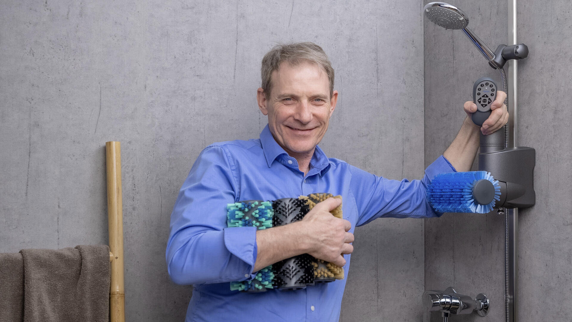 Ein lächelnder Mann in einem blauen Hemd steht in einer modernen Dusche, hält mehrere bunte Reinigungsbürsten in der Hand und hält mit der anderen Hand den Duschkopf fest. Ein braunes Handtuch hängt in der Nähe auf einem Bambusgestell an einer grauen Wand.