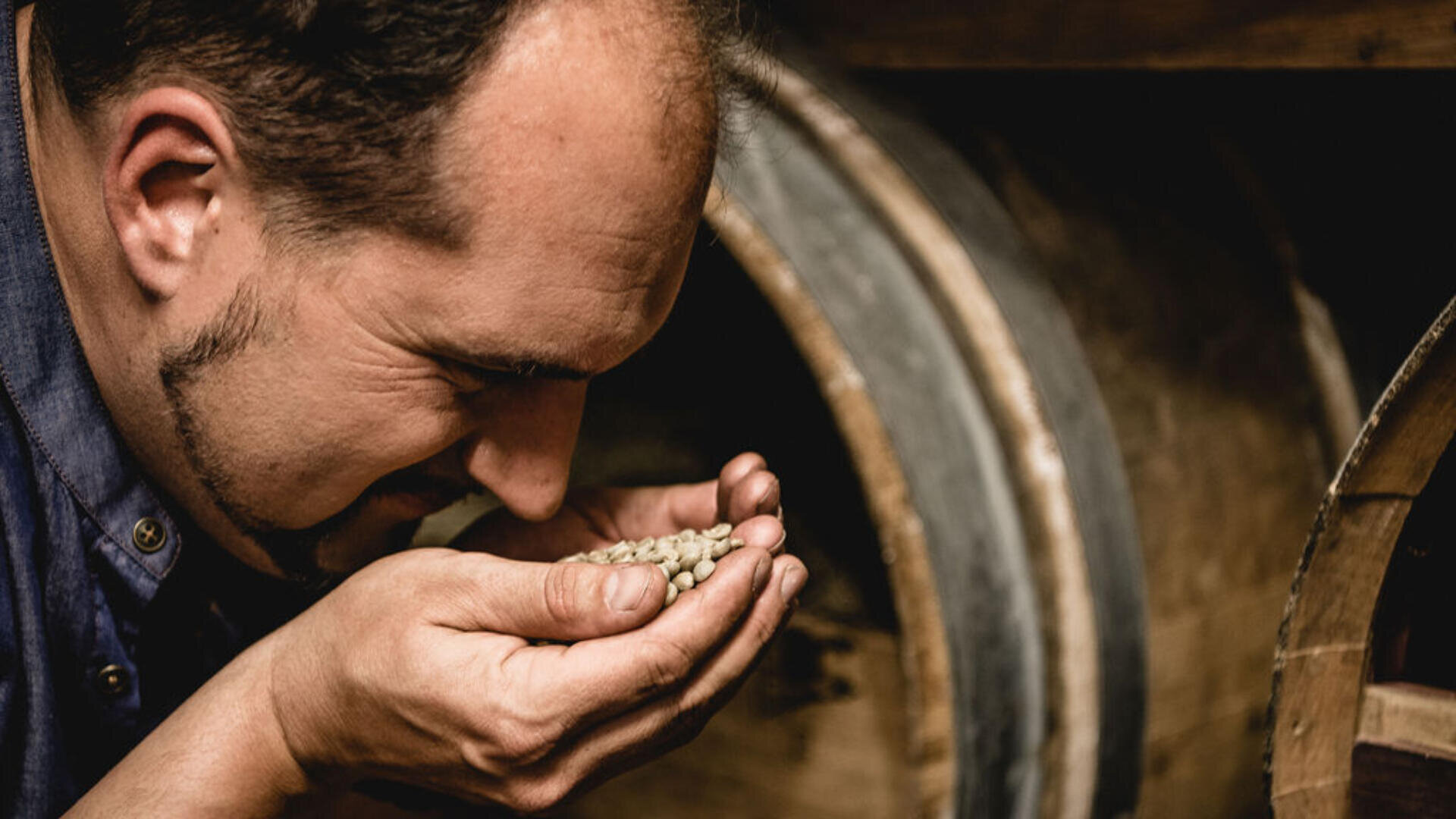 Ein Mann hält eine Handvoll grüner Kaffeebohnen in der Hand und riecht daran. Im Hintergrund sind Holzfässer zu sehen, die auf eine sorgfältige Prüfung der Qualität der Bohnen hindeuten.