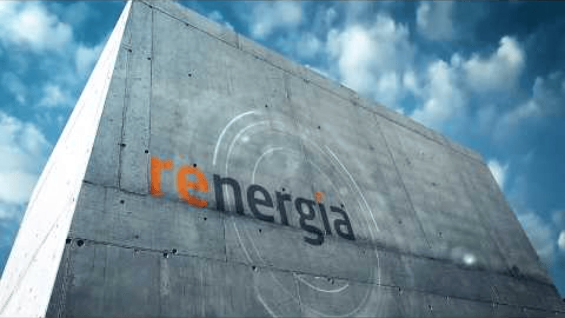 Eine Betonwand, auf der das Wort rēnergia steht, mit rē in Orange und nergia in Schwarz, unter einem blauen Himmel mit vereinzelten Wolken.