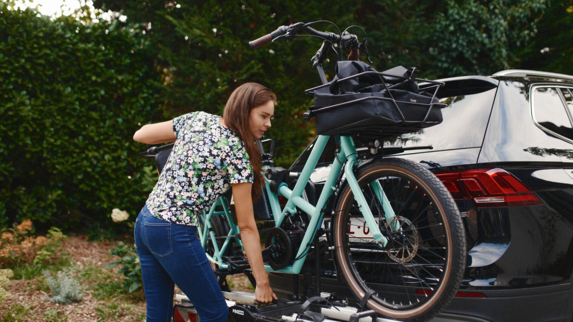 Eine Frau in geblümter Bluse und Jeans lädt ein hellblaues Fahrrad auf einen Fahrradträger, der hinten an einem schwarzen Auto befestigt ist, während im Hintergrund Grünflächen zu sehen sind.