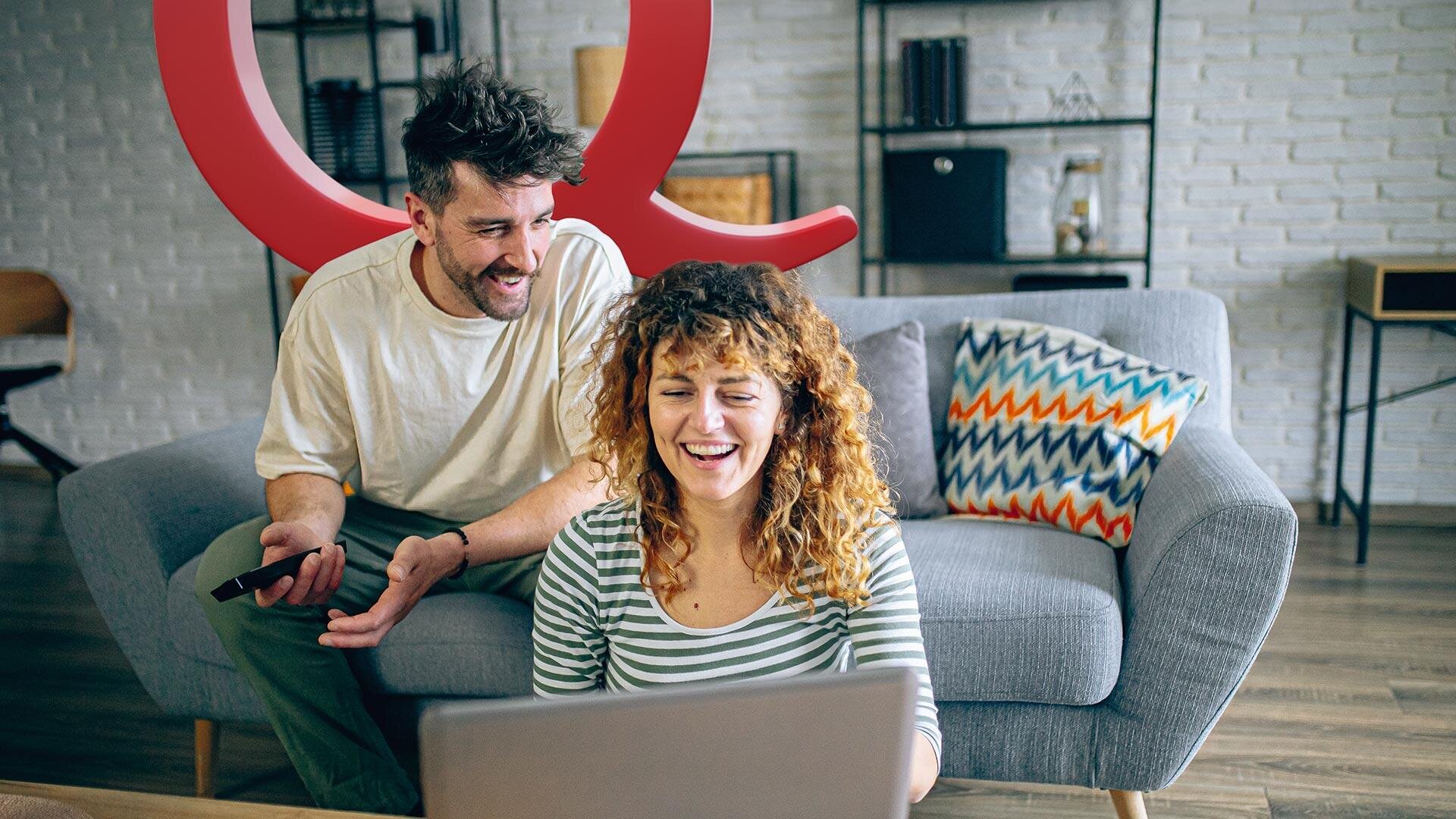 Eine lächelnde Frau sitzt auf einer Couch und benutzt einen Laptop, während ein Mann neben ihr spricht und gestikuliert. Beide wirken glücklich und entspannt in einem modernen Wohnzimmer mit bunten Kissen.