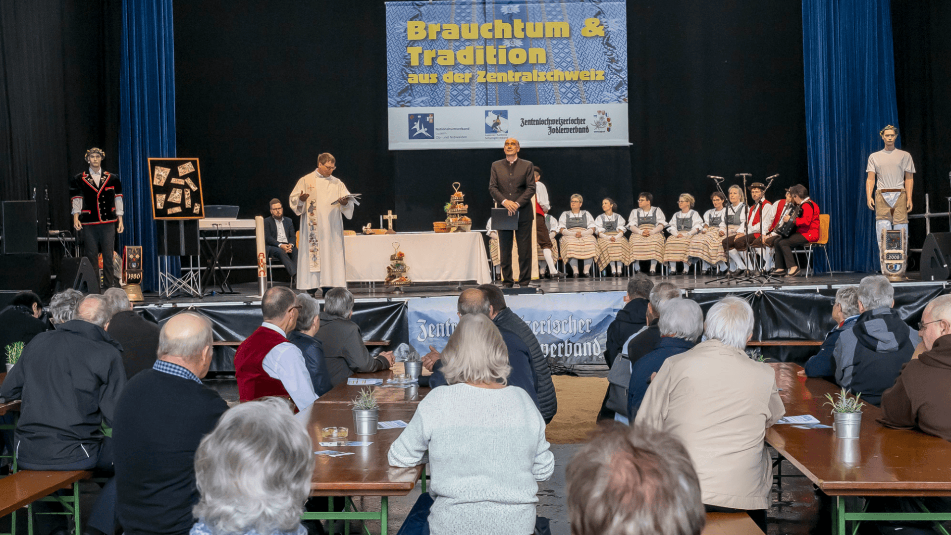 Eine kulturelle Veranstaltung auf der Bühne mit Menschen in traditioneller Schweizer Kleidung, einem Priester, der eine Rede hält, Musikern und kostümierten Puppen. Das Publikum sitzt an Tischen mit Blick auf die Bühne unter einem Transparent mit der Aufschrift "Brauchtum & Tradition aus der Zentralschweiz".