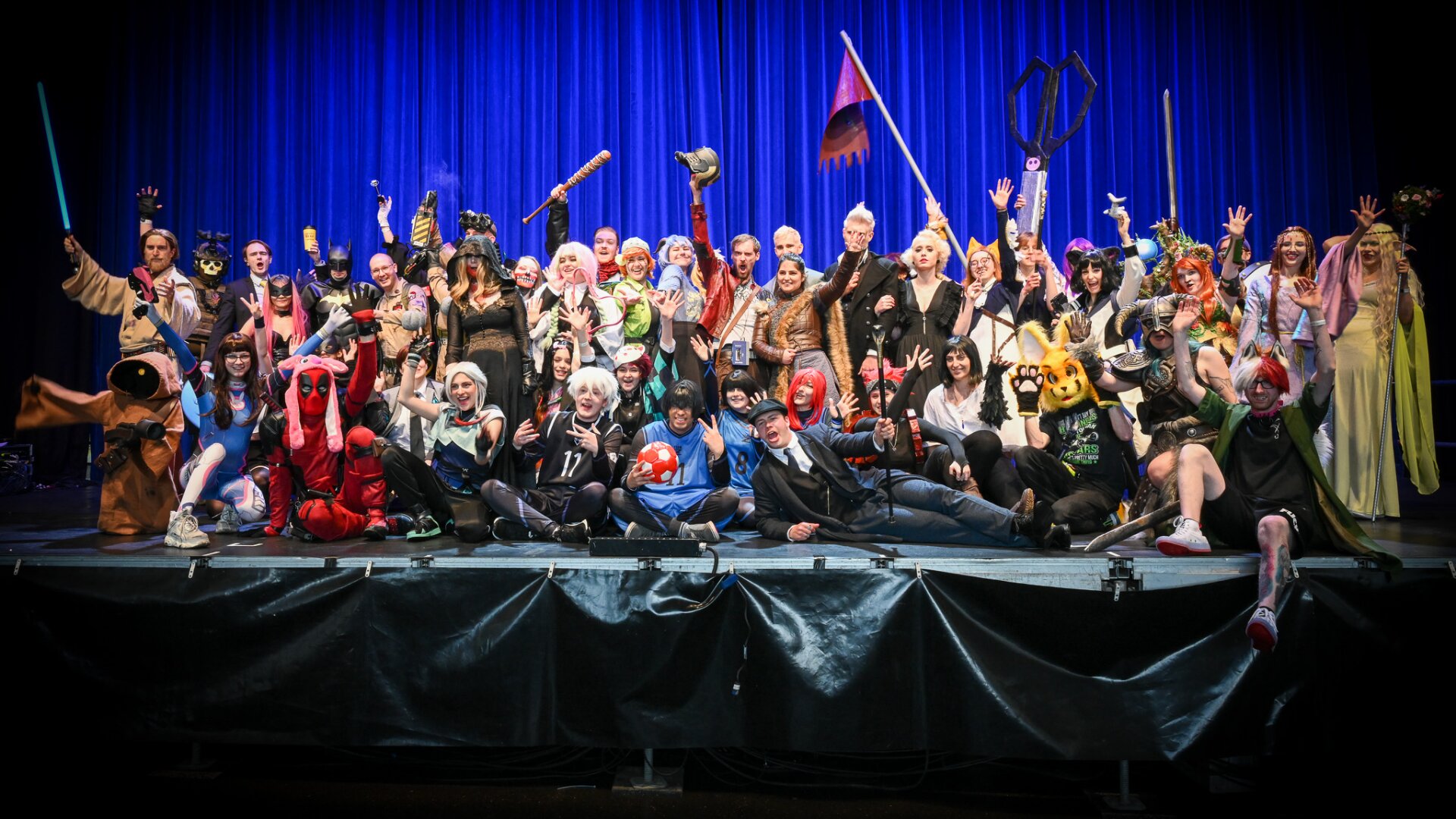 Eine große Gruppe von Menschen in farbenfrohen, aufwendigen Cosplay-Kostümen posiert begeistert auf der Bühne vor einem blauen Vorhang, hält verschiedene Requisiten und winkt in die Kamera.