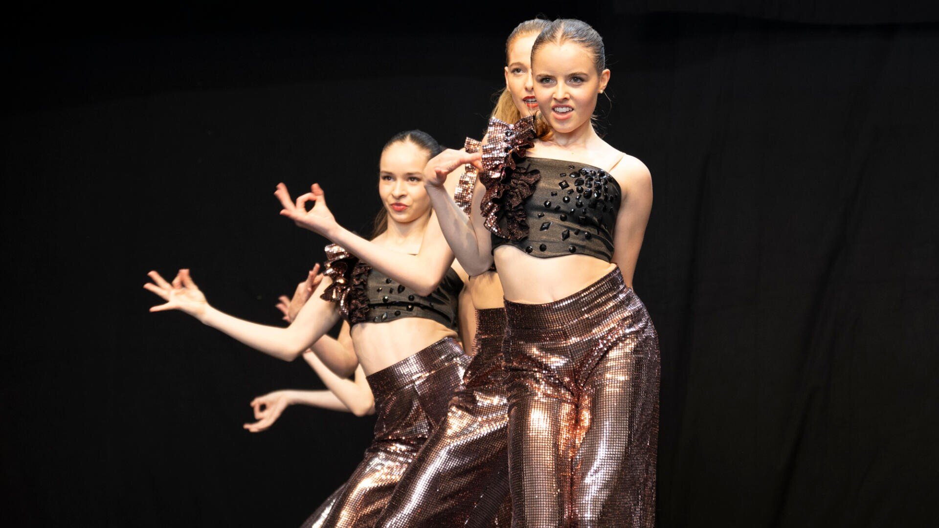 Drei junge Tänzerinnen in passenden bronzefarbenen Hosen und schwarzen Paillettenoberteilen führen auf der Bühne vor einem dunklen Hintergrund eine Choreografie auf. Der Haupttänzer steht vorne und macht eine dramatische Handbewegung.
