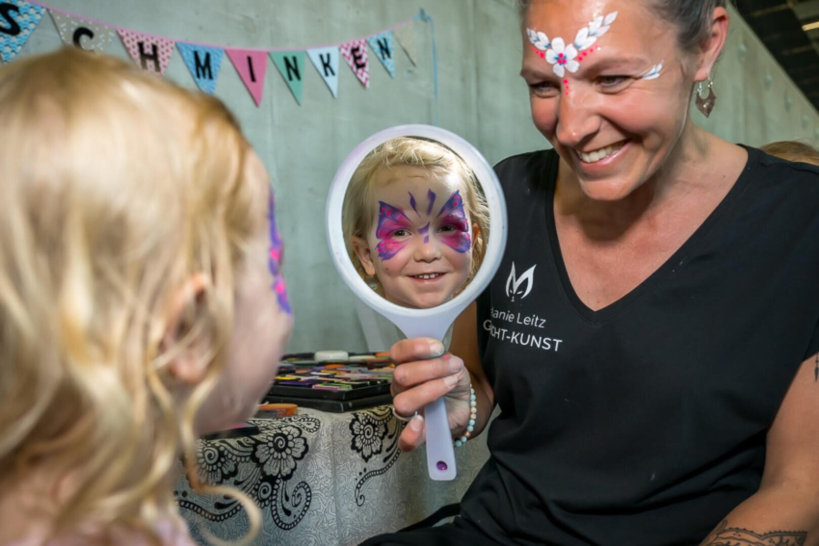 Eine lächelnde Frau hält einem jungen Mädchen mit buntem Schmetterlingsgesicht einen Spiegel vor, das sein Spiegelbild betrachtet. Beide haben bemalte Gesichter und stehen an einem Tisch mit Schminkutensilien. Im Hintergrund hängen Wimpel.