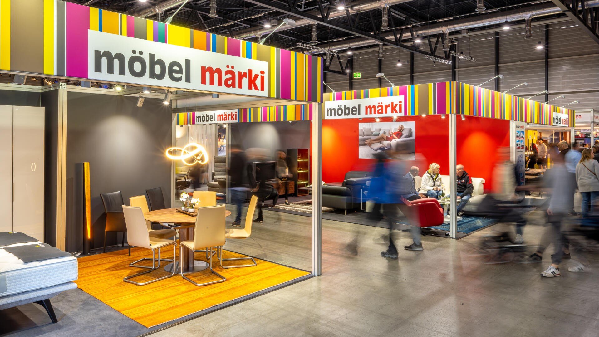 Ein belebter Möbel-Showroom mit bunten Schildern mit der Aufschrift möbel märki, auf denen verschiedene moderne Möbelstücke ausgestellt sind. Die Leute laufen herum und stöbern, und an einigen Ständen sind Esstische und Sofas ausgestellt.