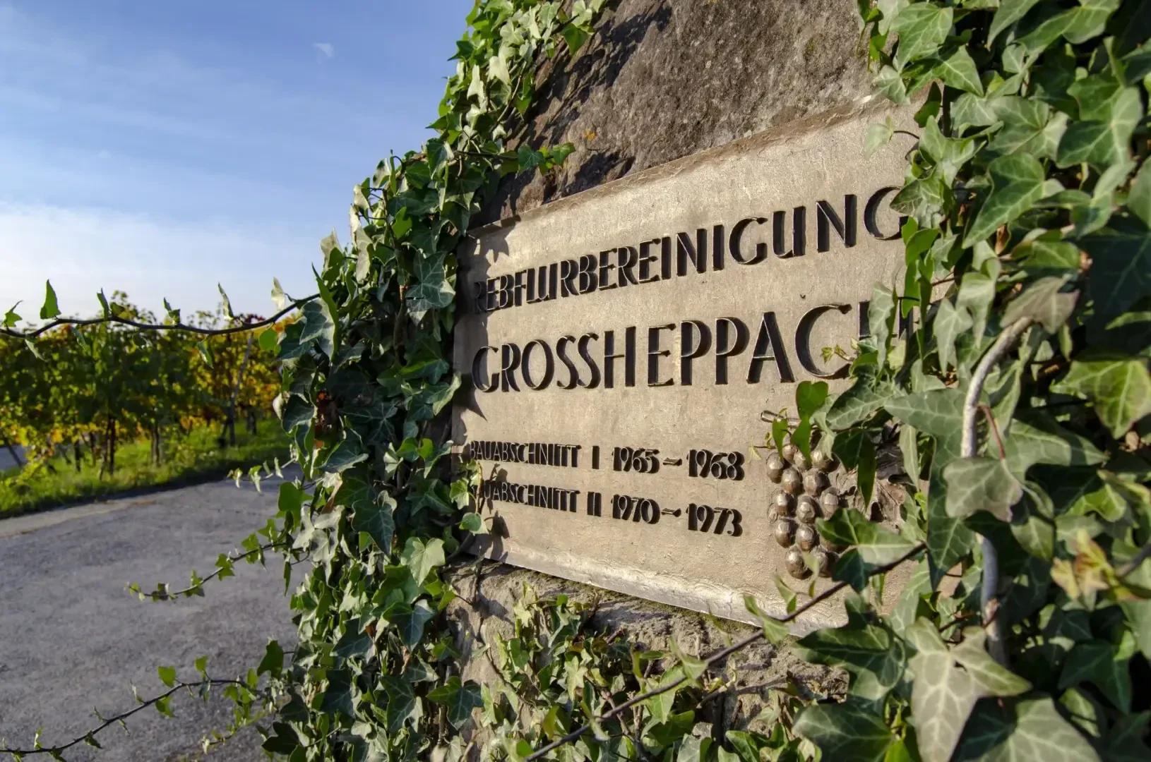 Auf einer Steintafel, die teilweise mit grünem Efeu bewachsen ist, steht GEBURVEREINIGUNG GROSSHEPPACH und sind die Jahre 1965-1968 und 1970-1973 aufgeführt. Im Hintergrund ist ein Weinberg unter blauem Himmel zu sehen.