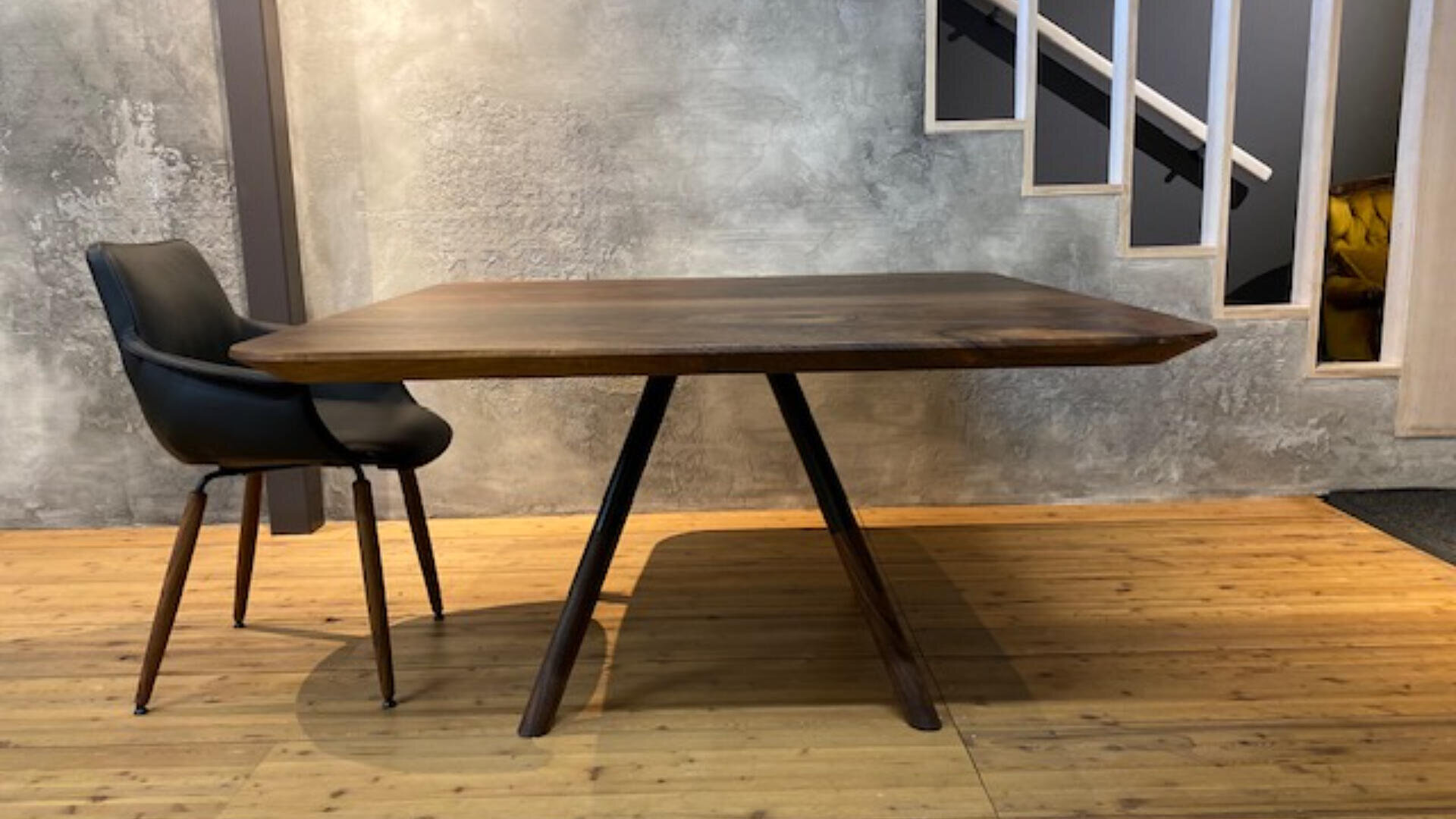 Ein moderner schwarzer Stuhl steht neben einem rechteckigen Holztisch mit abgewinkelten Beinen auf einem Holzboden, vor einer Wand mit Betonstruktur und einer Treppe mit weißem Geländer im Hintergrund.