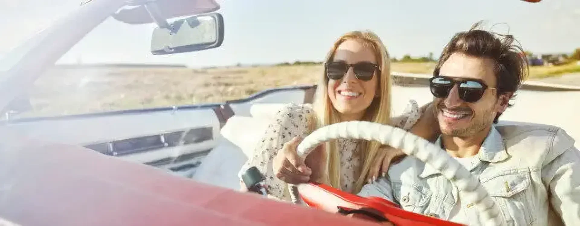 Ein lächelndes Paar mit Sonnenbrille sitzt an einem sonnigen Tag in einem Cabrio mit offenem Verdeck. Die Frau zeigt nach vorne, während der Mann fährt, beide genießen den Moment.