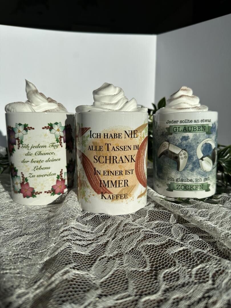 Drei dekorierte, mit Schlagsahne gefüllte Becher mit deutschem Text und unterschiedlichen Designs: Blumenmuster, orangefarbener Hintergrund mit Kaffee-Zitat und Gaming-Thema mit Game-Controller und Kopfhörer.