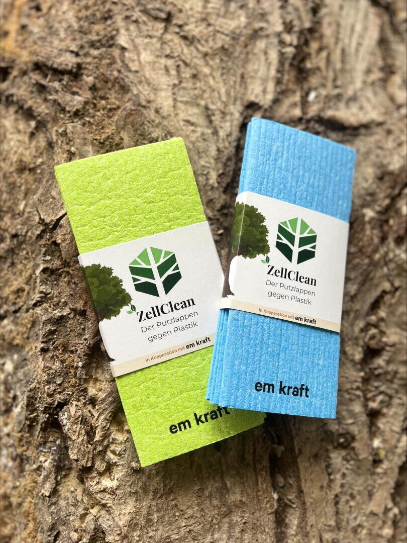Zwei umweltfreundliche ZellClean-Reinigungstücher, ein grünes und ein blaues, ruhen auf strukturierter Baumrinde. Jedes Tuch hat eine Papierbanderole mit einem grünen Baumlogo und einem Text, der für eine plastikfreie Reinigung wirbt.