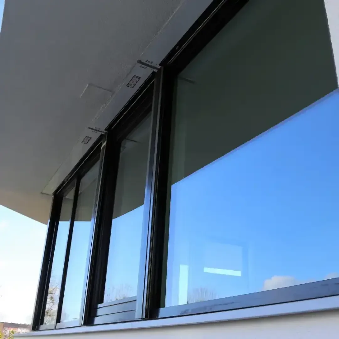 Nahaufnahme eines modernen Schiebefensters mit schwarzem Rahmen, in dem sich der blaue Himmel und ein Teil der Außenfassade eines Gebäudes spiegeln.