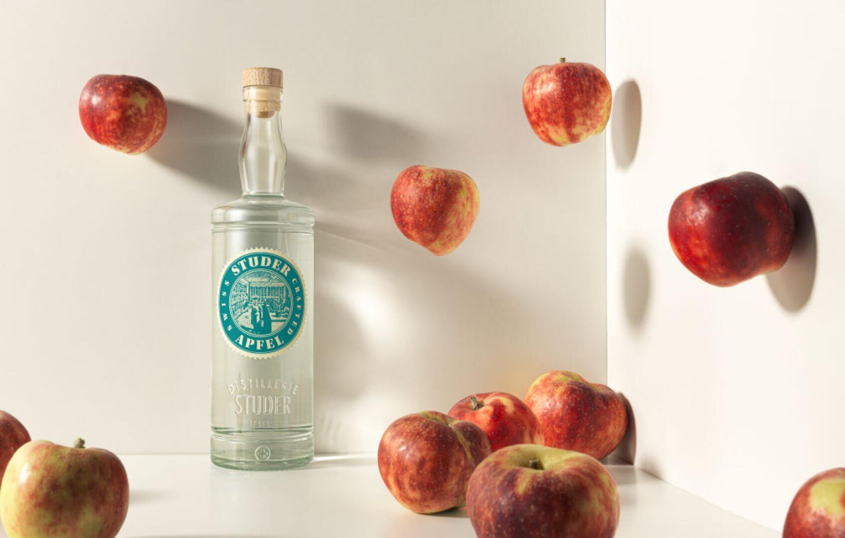Eine mit einem Korken versehene Klarglasflasche mit Studer-Apfel-Likör steht aufrecht, umgeben von frischen roten Äpfeln, von denen einige auf der Oberfläche ruhen und andere vor einem weißen Hintergrund zu schweben scheinen.