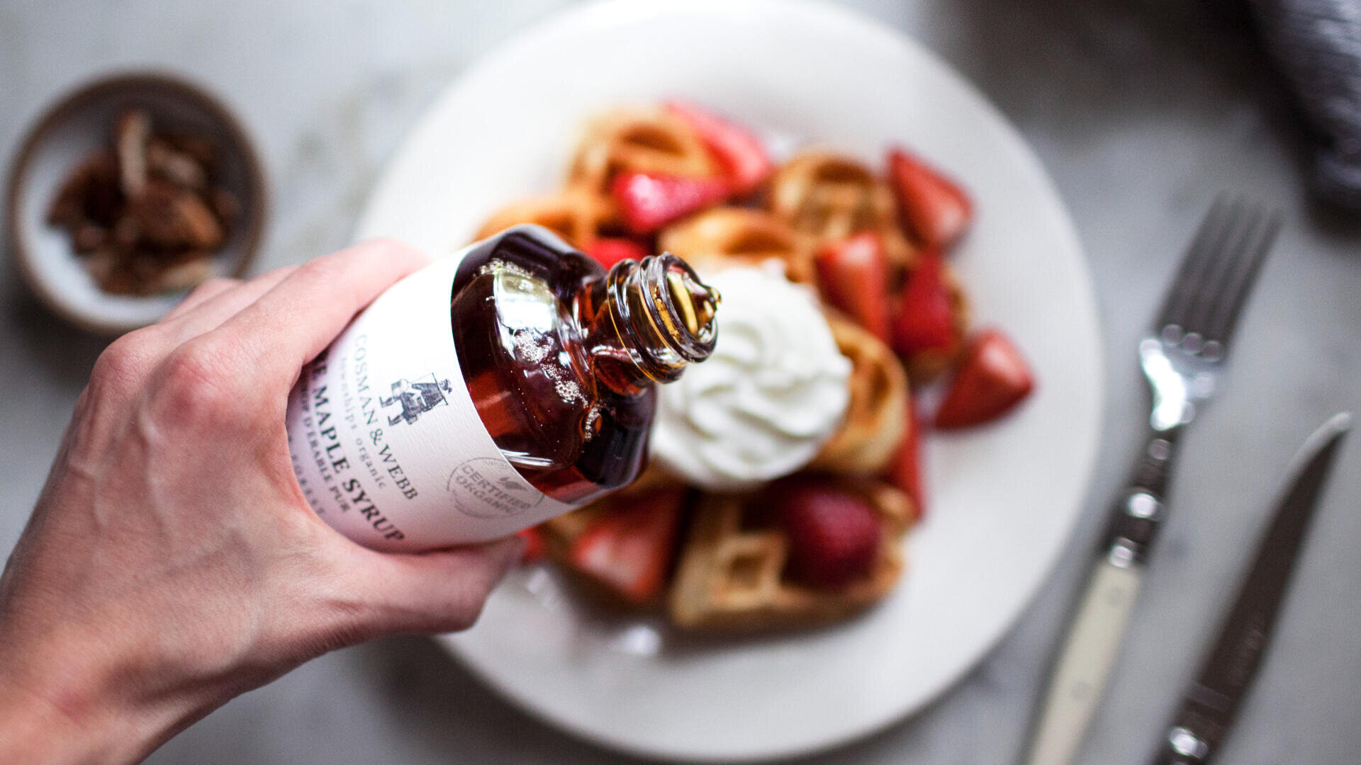 Eine Hand gießt Ahornsirup aus einer Glasflasche auf einen Teller mit Waffeln, die mit Schlagsahne und Erdbeeren belegt sind; eine Gabel, ein Messer und eine kleine Schale mit Pekannüssen stehen daneben.
