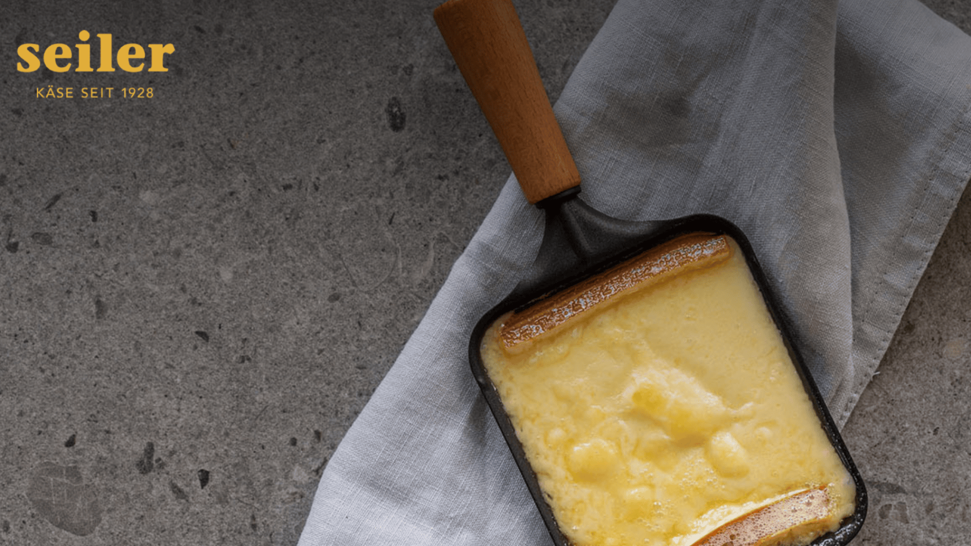 Eine kleine schwarze Pfanne mit geschmolzenem Käse steht auf einem hellgrauen Tuch auf einer Steinfläche. In der oberen linken Ecke steht in gelber Schrift "Seiler KÄSE SEIT 1928".