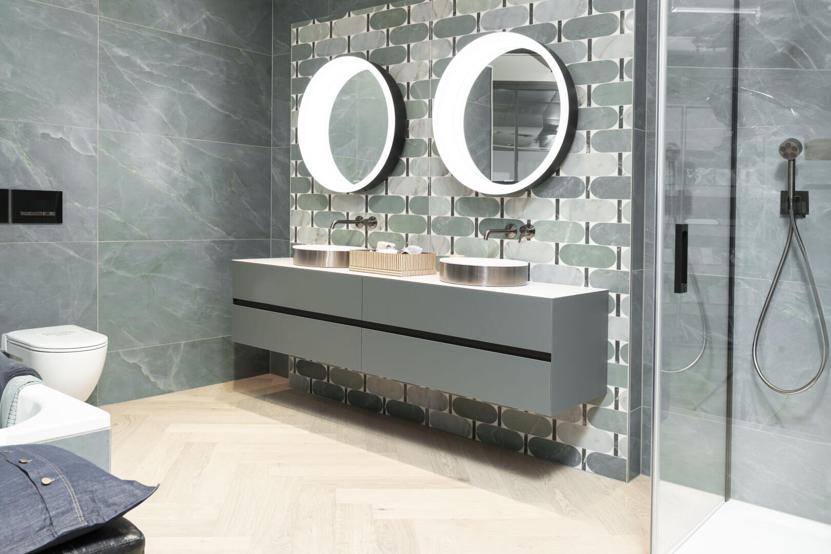 Modernes Badezimmer mit grün gefliesten Wänden, doppeltem, schwebendem Waschtisch mit zwei Gefäßwaschbecken, runden Spiegeln mit Hintergrundbeleuchtung, einer Glasduschabtrennung und einer Toilette. Der Fischgrätboden aus hellem Holz verleiht dem Raum eine warme Atmosphäre.