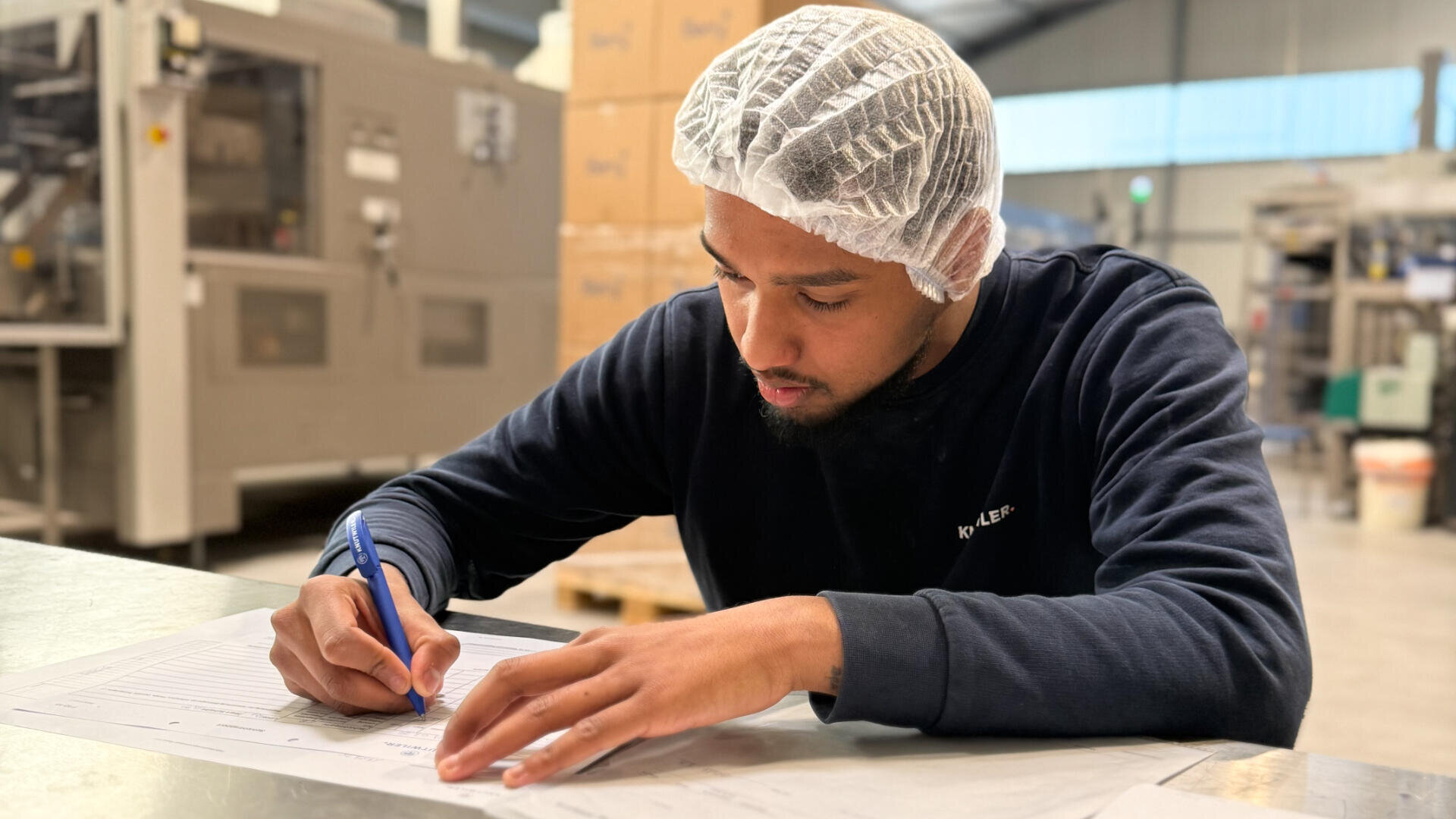 Ein Mann mit Haarnetz und marineblauem Pullover sitzt an einem Tisch in einer Fabrik und füllt konzentriert Papierkram mit einem blauen Stift aus. Im Hintergrund sind Industrieanlagen und gestapelte Kisten zu sehen.