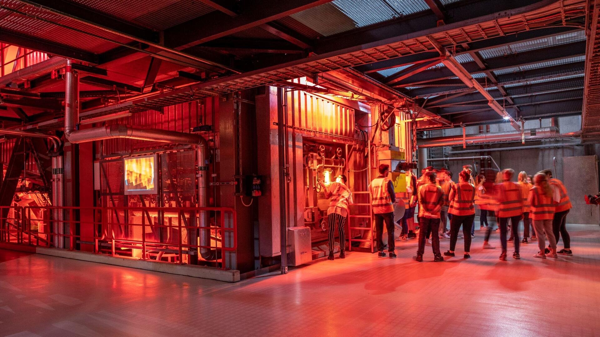 Eine Gruppe von Personen mit orangefarbenen Sicherheitswesten steht in einer rot beleuchteten Industrieanlage und beobachtet Maschinen und Arbeiter durch Sicherheitsschranken.
