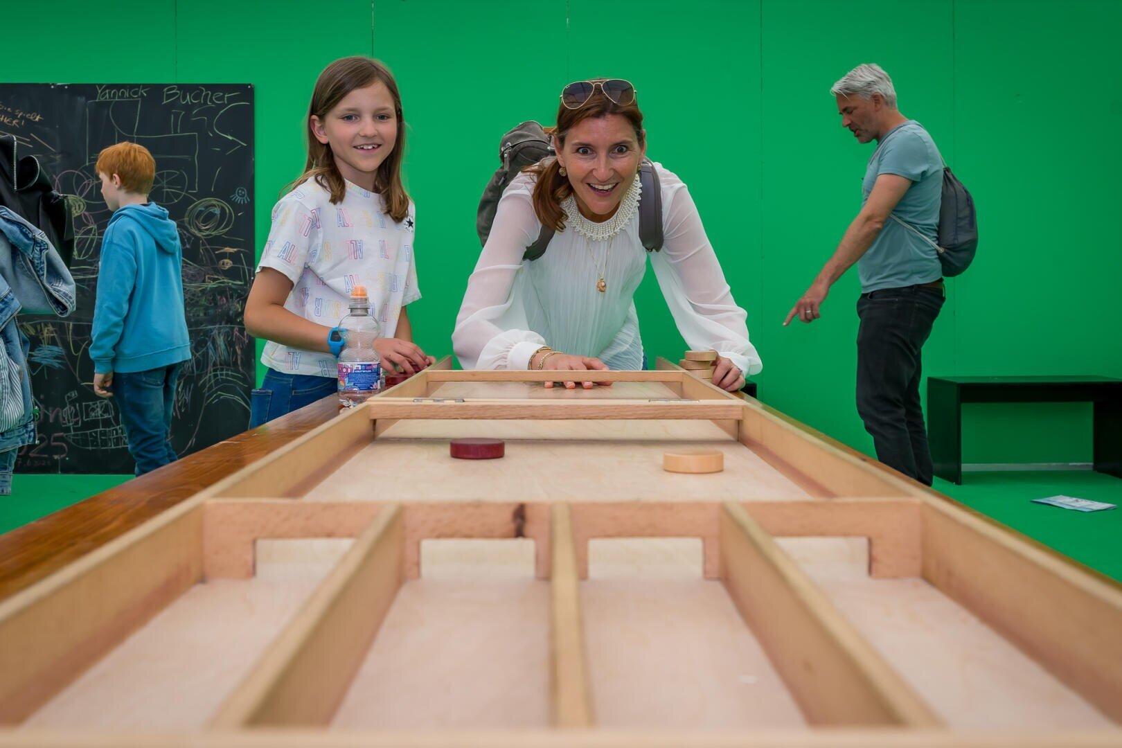 Eine Frau und ein Mädchen lächeln, während sie ein Tischspiel auf einem Holzbrett spielen. Im Hintergrund sind zwei Personen zu sehen, von denen eine auf eine Kreidetafel an der Wand schreibt und eine andere in der Nähe steht. Die Szene spielt sich vor einem grünen Hintergrund ab.