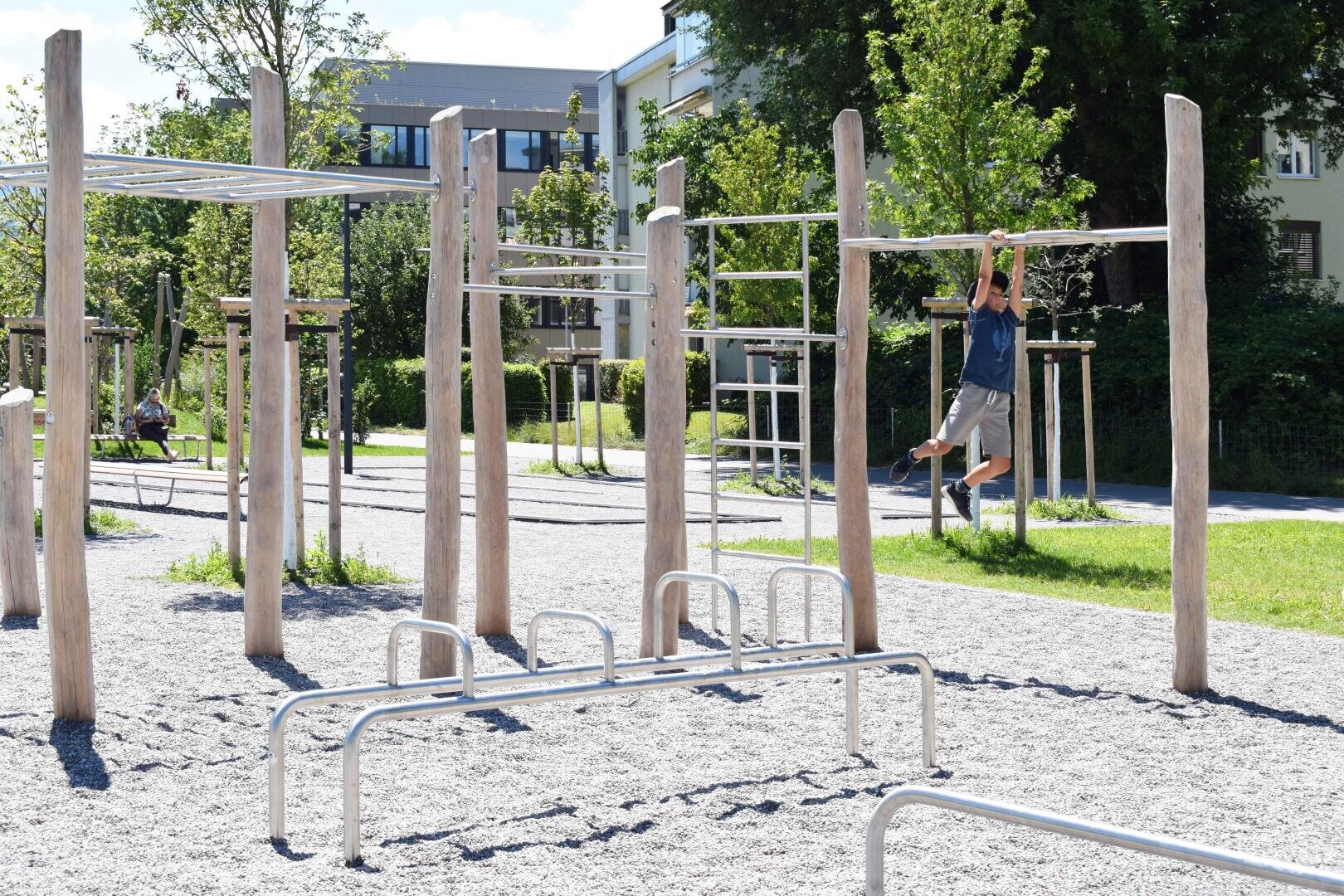 Ein Kind hängt an einem sonnigen Tag an einem Klettergerüst auf einem Spielplatz mit Holzgeräten im Freien. Im Hintergrund sind Gras, Bäume und nahe gelegene Gebäude zu sehen.