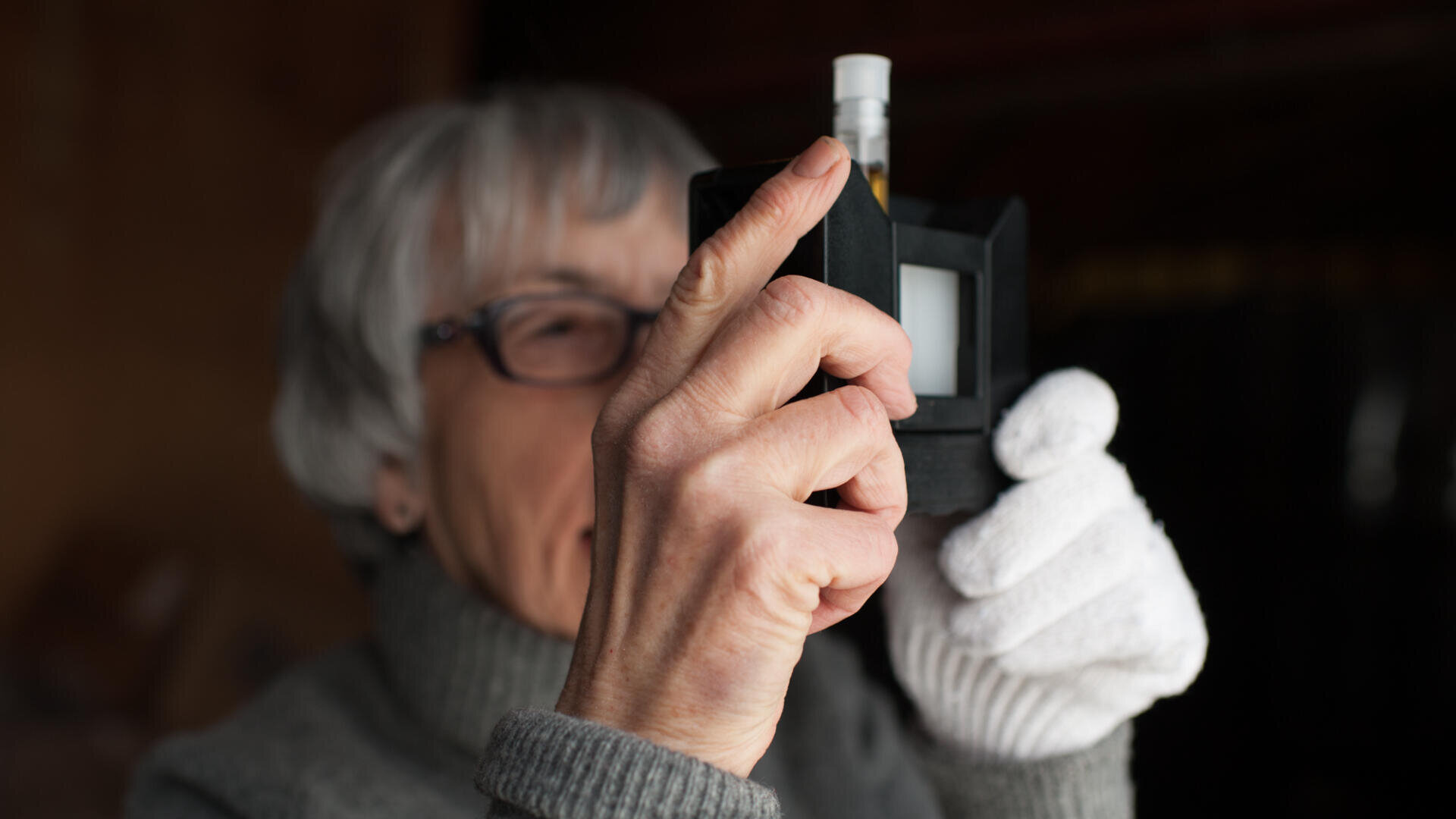 Eine ältere Person mit Brille und weißen Handschuhen betrachtet ein fotografisches Dia mit einem Diabetrachter in der Hand.