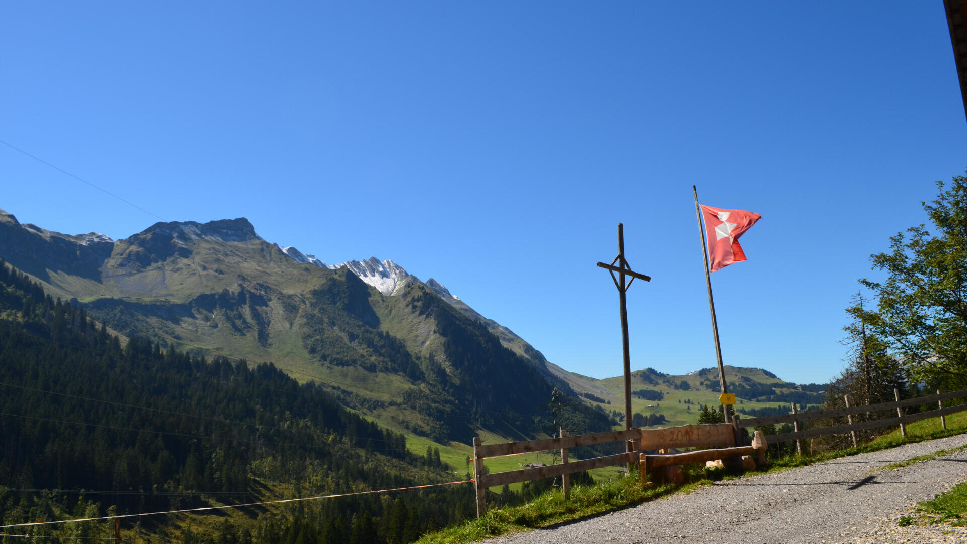 Eine Schweizer Fahne auf einer Stange und ein Holzkreuz stehen neben einem rustikalen Zaun mit Blick auf grüne Hügel, Kiefernwälder und ferne felsige Berge unter einem klaren blauen Himmel.