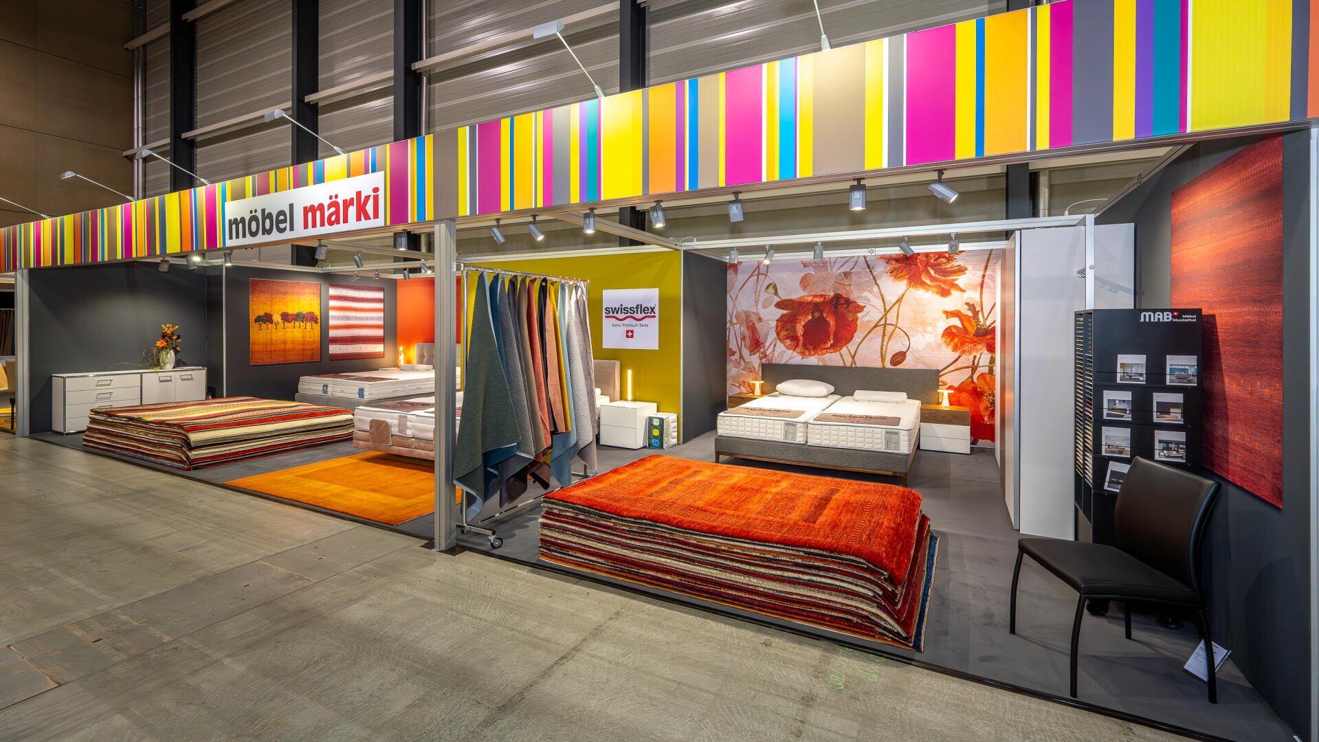 In einem farbenfrohen Möbelhaus stehen Betten mit Matratzen, gestapelte Teppiche, ein Stuhl und Wandkunst unter einem gelb-orange gestreiften Schild mit der Aufschrift möbel märki. Der Stand ist gut beleuchtet und modern.