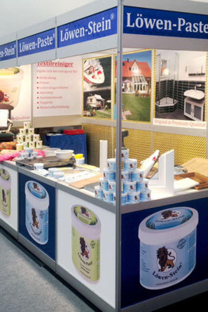 Auf einem Messestand von Löwen-Paste stehen gestapelte Dosen Reinigungspaste mit einem Löwen-Logo, Informationsplakate und Broschüren auf einem Tresen. Über dem Stand sind Banner mit dem Markennamen zu sehen.