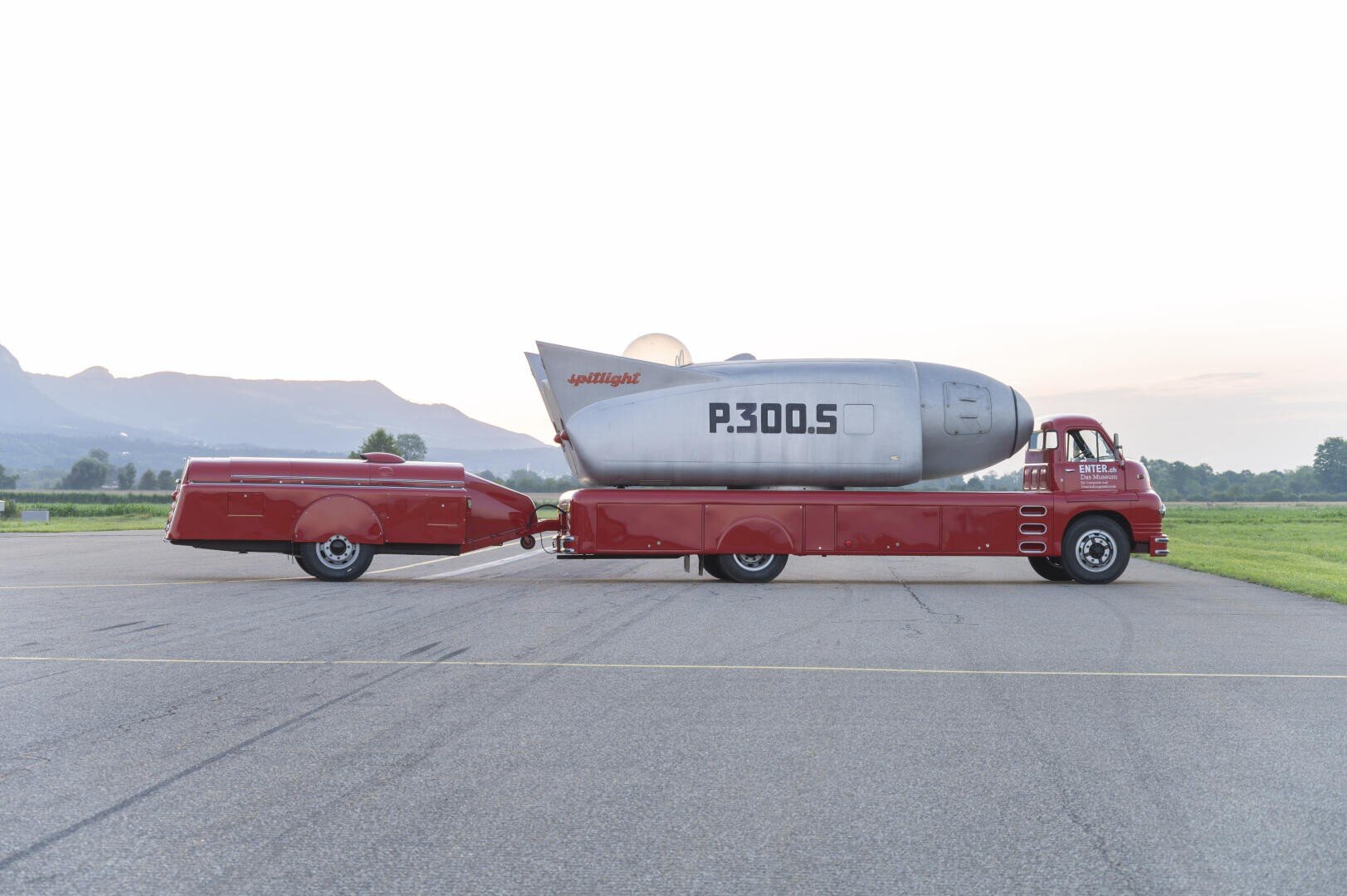 Ein roter Oldtimer-Lkw transportiert ein großes silbernes raketenförmiges Fahrzeug mit der Aufschrift P.300.S auf einer Pritsche, das einen passenden roten stromlinienförmigen Anhänger zieht, der auf einem Rollfeld mit Bergen im Hintergrund geparkt ist.
