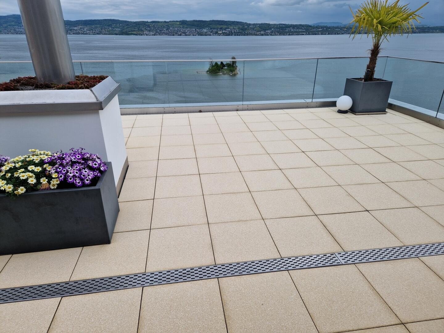 Eine geflieste Dachterrasse mit Topfblumen und einer Palme bietet einen Blick auf einen großen See und ferne grüne Hügel unter einem bewölkten Himmel. Ein Glasgeländer säumt den Rand der Terrasse.