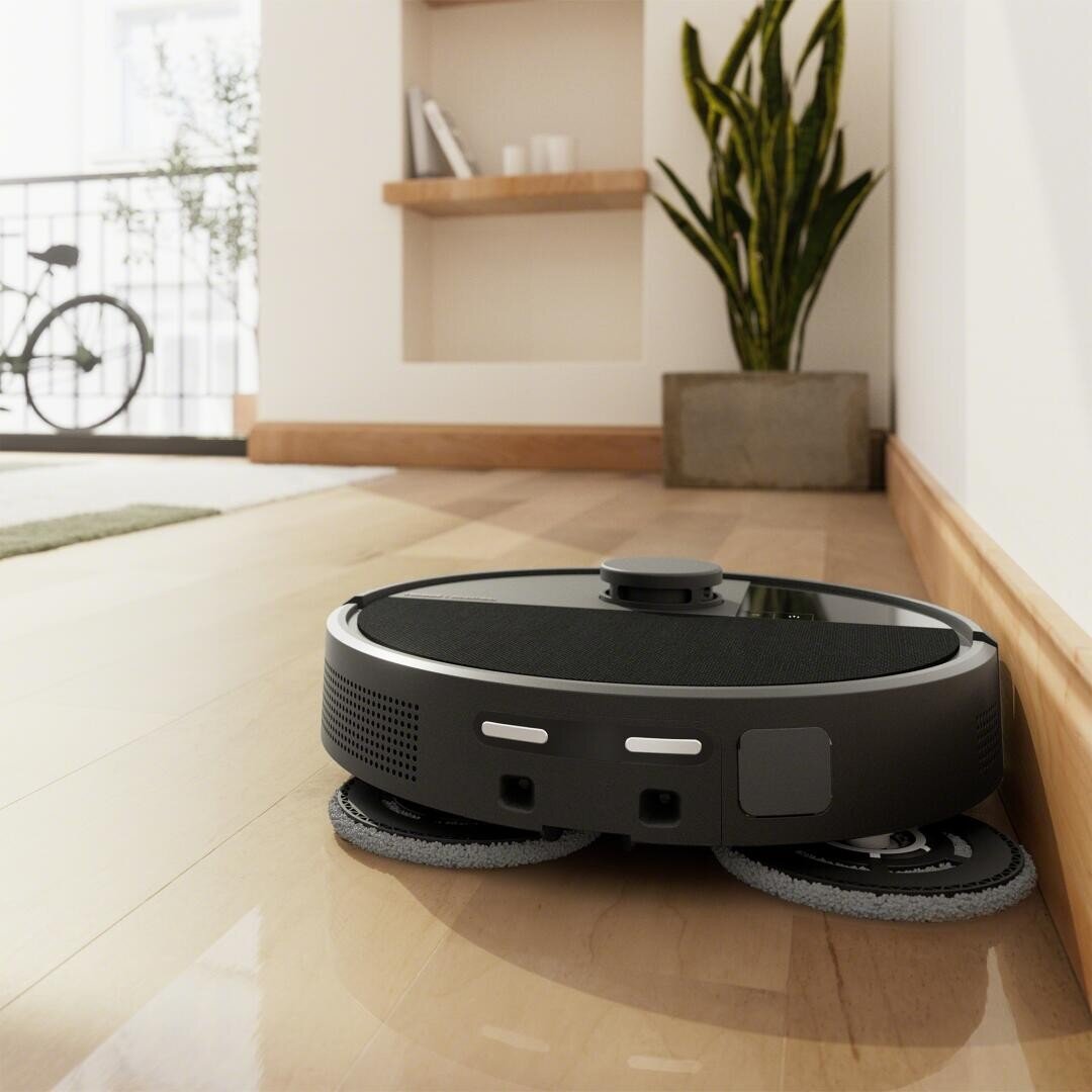 Ein Staubsaugerroboter mit rotierenden Bürsten reinigt einen glänzenden Holzboden in der Nähe einer Wand in einer modernen Wohnung, mit einer Pflanze und Regalen im Hintergrund.