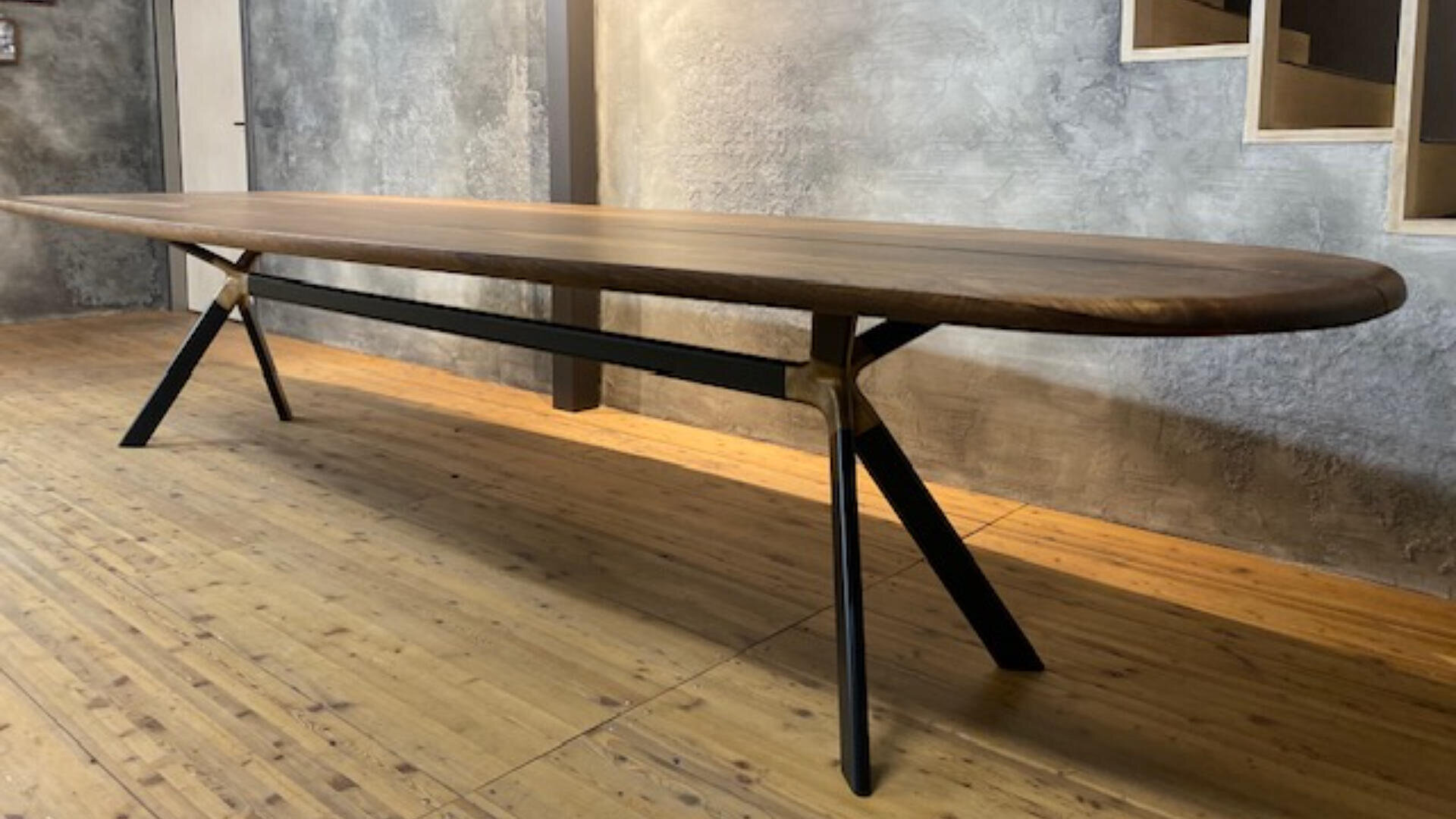 Ein langer, ovaler Holztisch mit schwarzen Metallbeinen steht auf einem hellen Holzboden vor einer strukturierten grauen Wand in einem modernen, minimalistischen Raum.