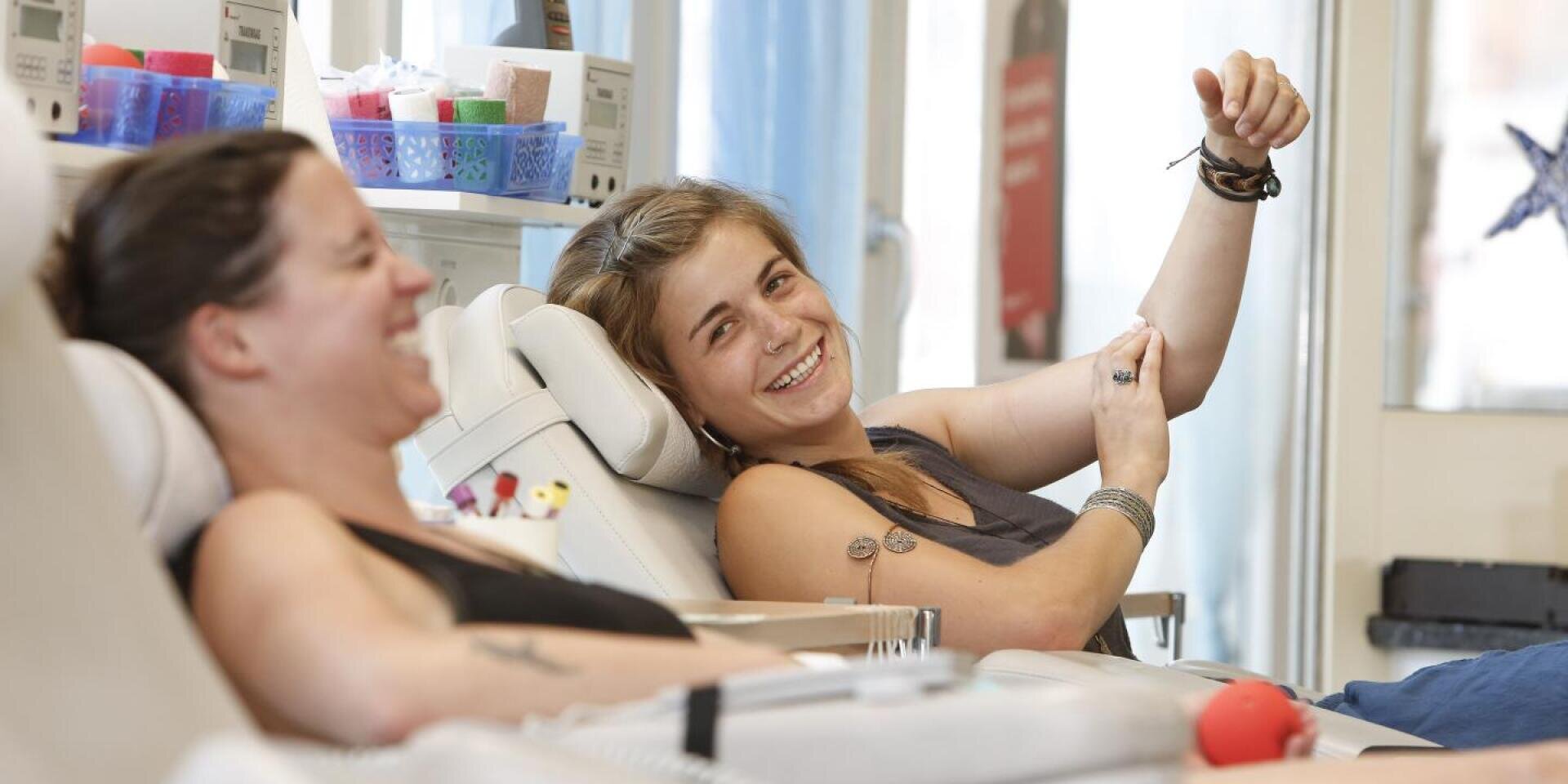 Zwei Frauen entspannen sich und lächeln, während sie Blut spenden, eine von ihnen hält ihren Arm hoch und schaut in die Kamera. Im Hintergrund sind medizinische Geräte und Zubehör zu sehen.