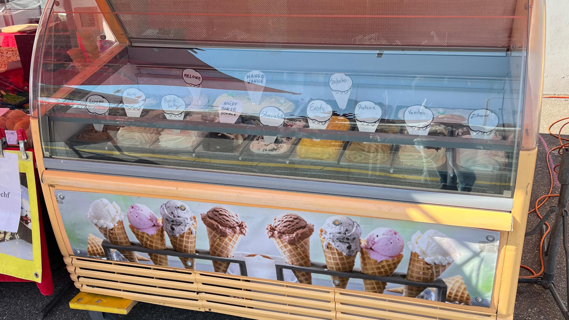 In einer Vitrine auf einem Markt im Freien werden verschiedene Becher mit buntem Eis und handgeschriebenen Geschmacksetiketten ausgestellt. Die Vitrine ist am unteren Rand mit Bildern von Eistüten verziert.