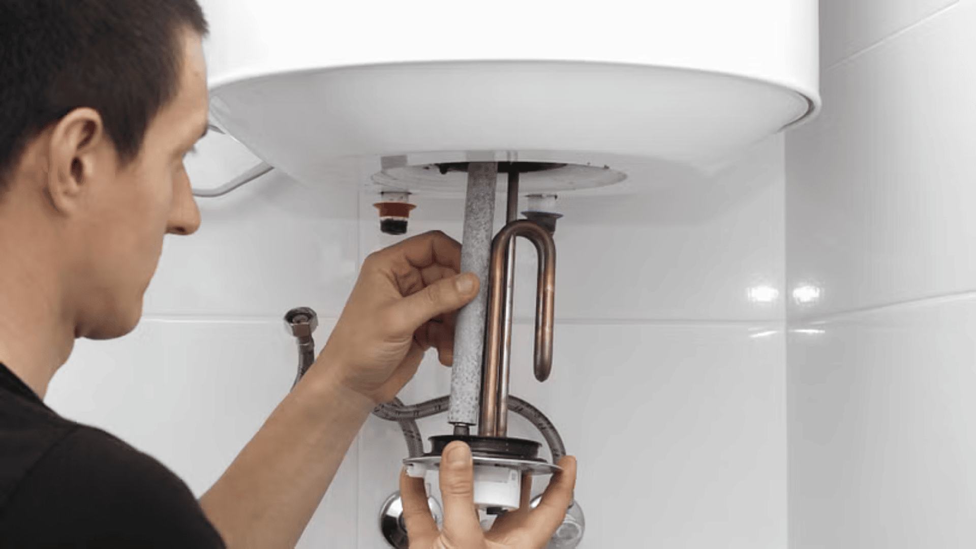 Eine Person repariert einen Warmwasserbereiter, indem sie dessen Heizelement aus dem an einer weiß gekachelten Wand montierten Gerät herausnimmt.