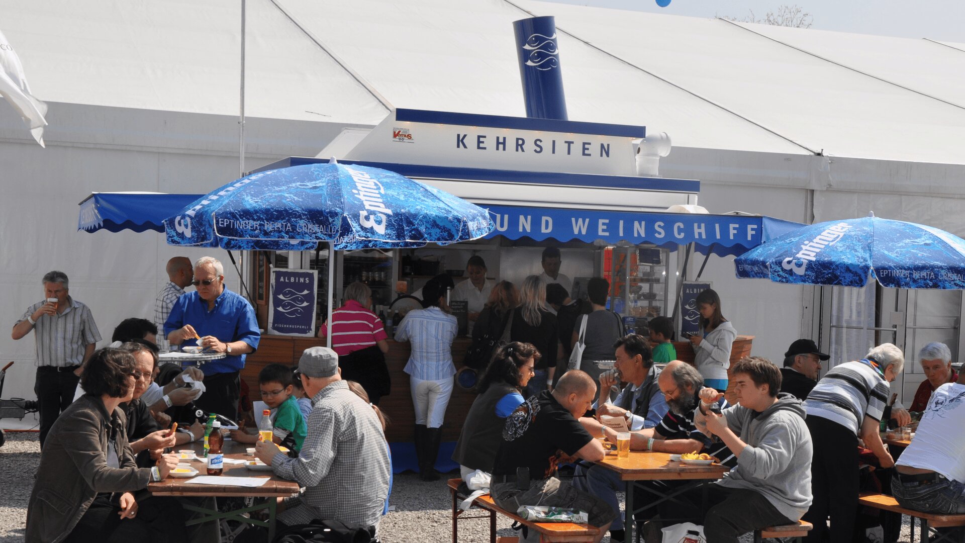 Die Menschen sitzen an Tischen im Freien, essen und trinken in der Nähe eines weißen Zeltes und eines Essensstandes mit der Aufschrift Kehrsiten. Einige stehen in der Schlange an der Theke; blaue Sonnenschirme spenden Schatten über den Tischen.