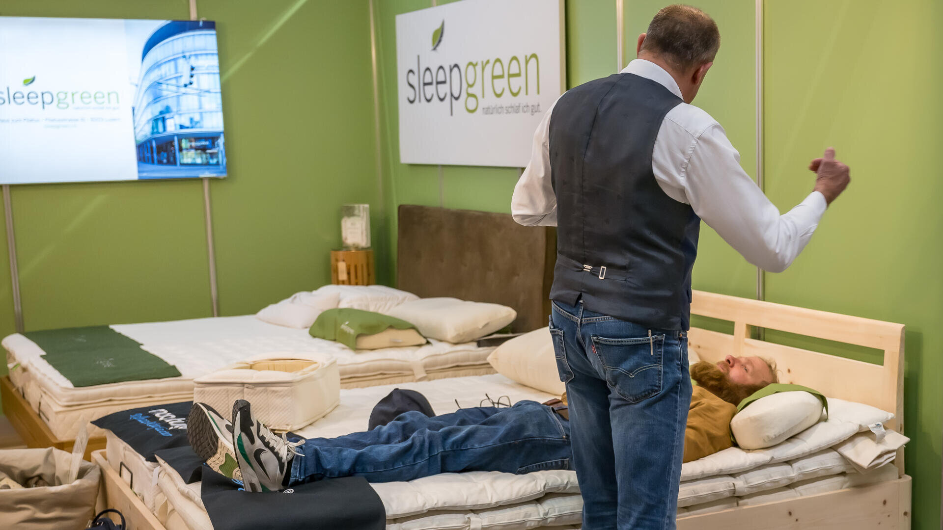 Ein Mann in einer Weste gestikuliert, während er neben einem anderen Mann steht, der auf einem Bett vor einer Sleepgreen-Matratzenausstellung in einem Ausstellungsraum mit grünen Wänden und Bettwaren liegt.