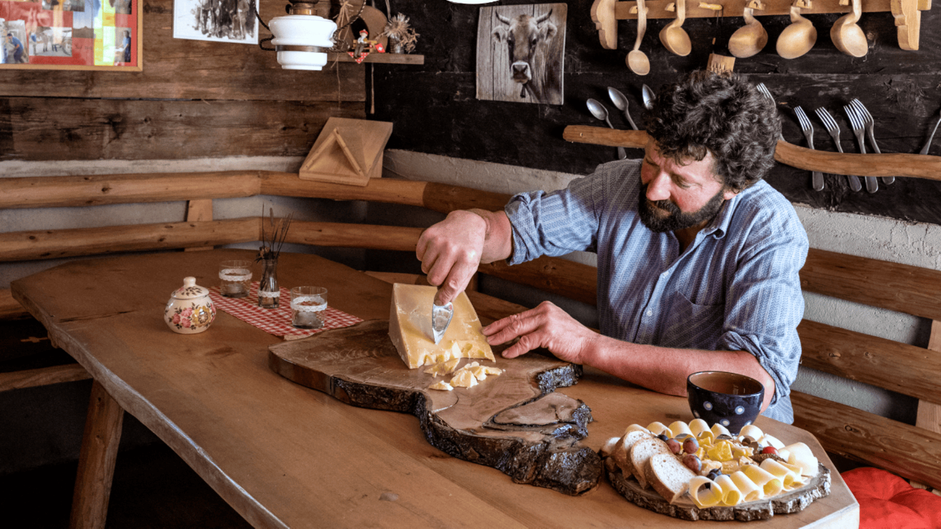 Ein Mann sitzt an einem rustikalen Holztisch in einer gemütlichen Hütte und schneidet Käse auf einem Holzbrett. Der Tisch ist mit einer Tasse, Brötchen und Käse gedeckt; Holzutensilien hängen an der Wand hinter ihm.
