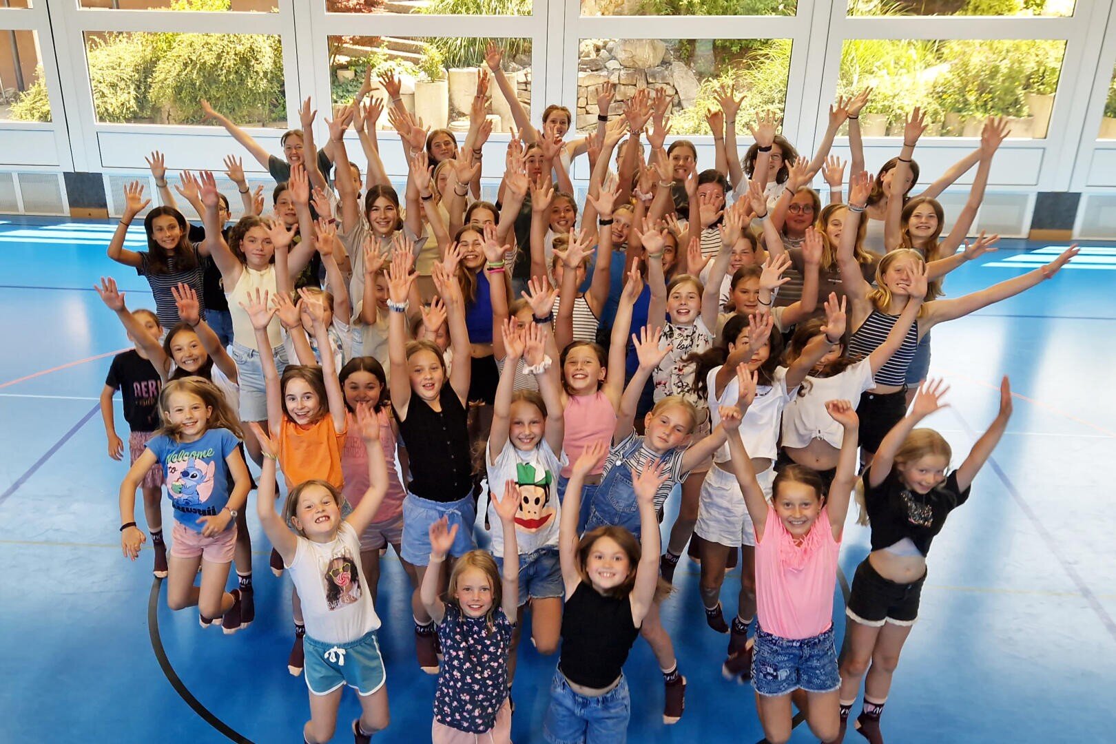 Eine große Gruppe lächelnder Kinder und einige Erwachsene stehen in einer Turnhalle mit blauem Boden und heben begeistert ihre Arme in Richtung Kamera. Im Hintergrund sind helle Fenster und Grünflächen zu sehen.
