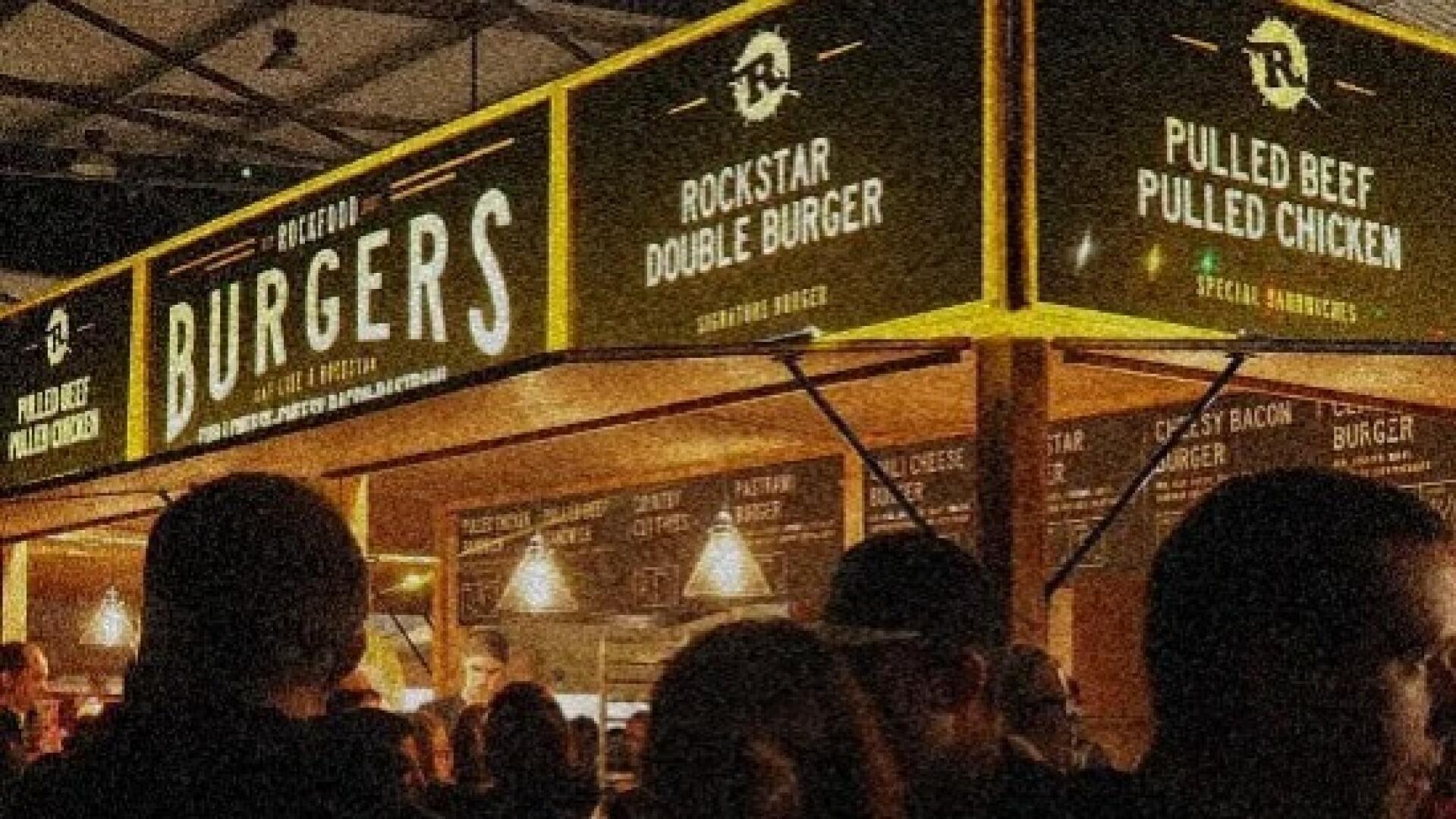 Ein belebter Burgerstand bei Nacht, mit beleuchteten Schildern, auf denen Speisekarten wie Rockstar Double Burger, Pulled Beef und Pulled Chicken stehen. Vor der Theke haben sich Menschenmassen versammelt.