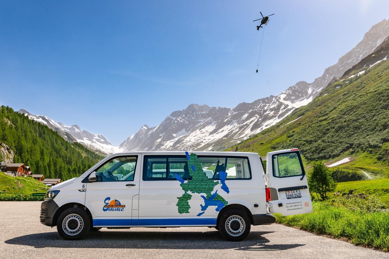 Ein weißer Lieferwagen mit einem Bergkartenaufkleber parkt auf einer Panoramastraße in einem grünen Alpental, mit schneebedeckten Gipfeln im Hintergrund. Darüber fliegt ein Hubschrauber, der eine an einem Kabel aufgehängte Last trägt.