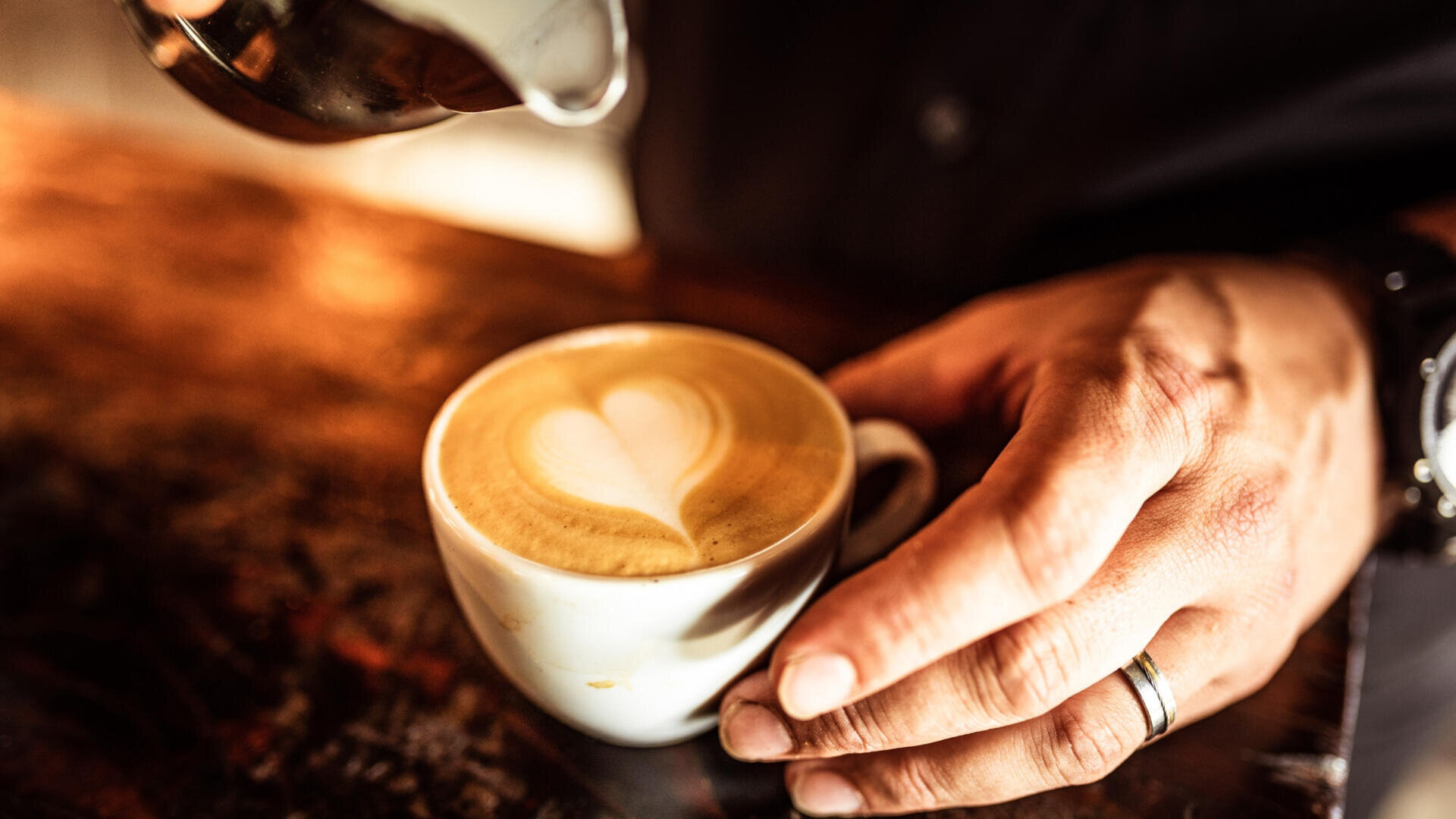 Eine Person mit einem silbernen Ring hält eine Tasse Kaffee mit einem herzförmigen Latte Art Design, während sie Milch in die Tasse auf einem Holztisch gießt.