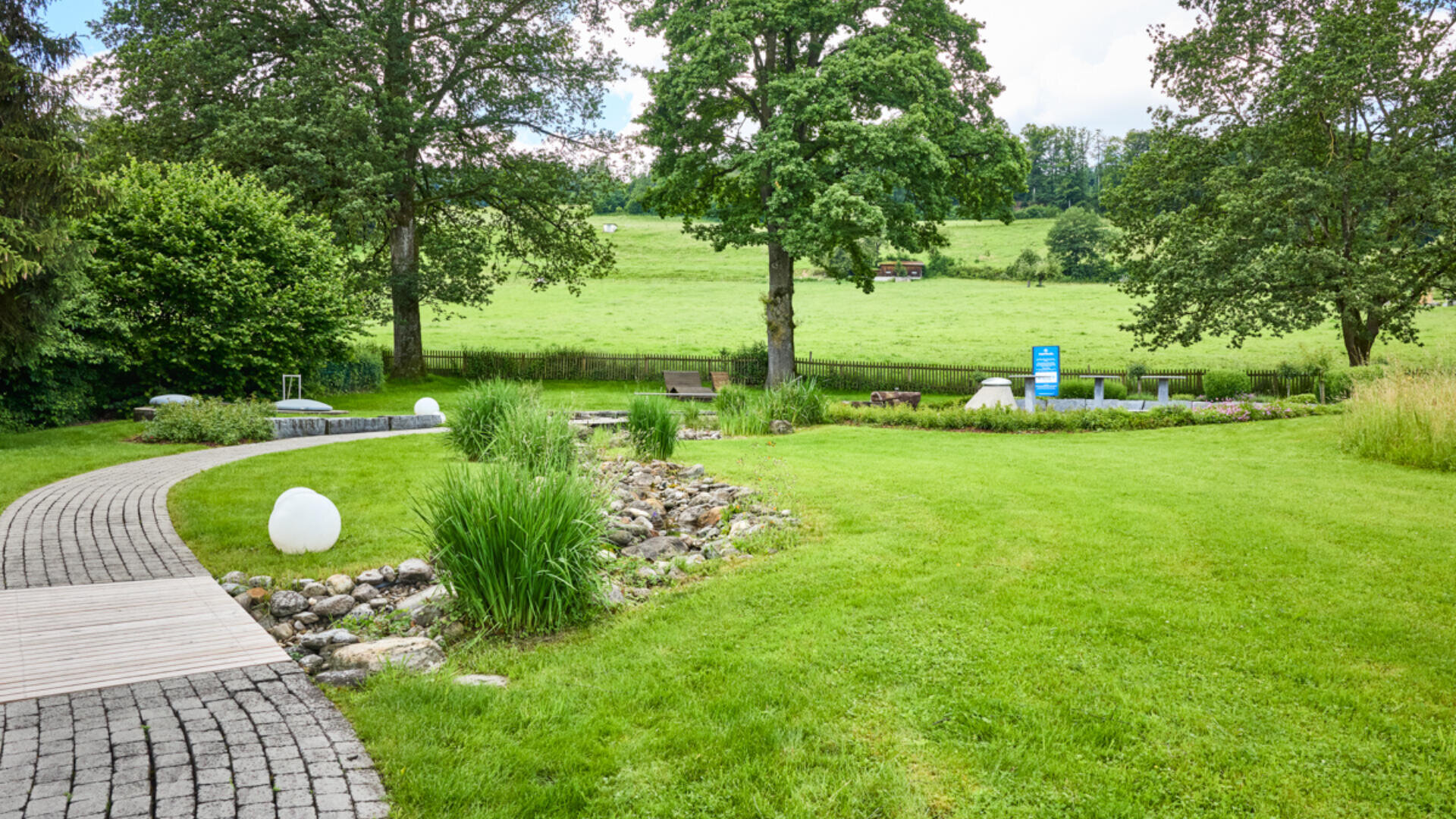 Ein Landschaftsgarten mit einem geschwungenen Steinweg, einem kleinen Bach, dekorativen weißen Kugeln, Bänken und Picknicktischen, umgeben von grünem Gras, Bäumen und einem offenen Feld im Hintergrund.