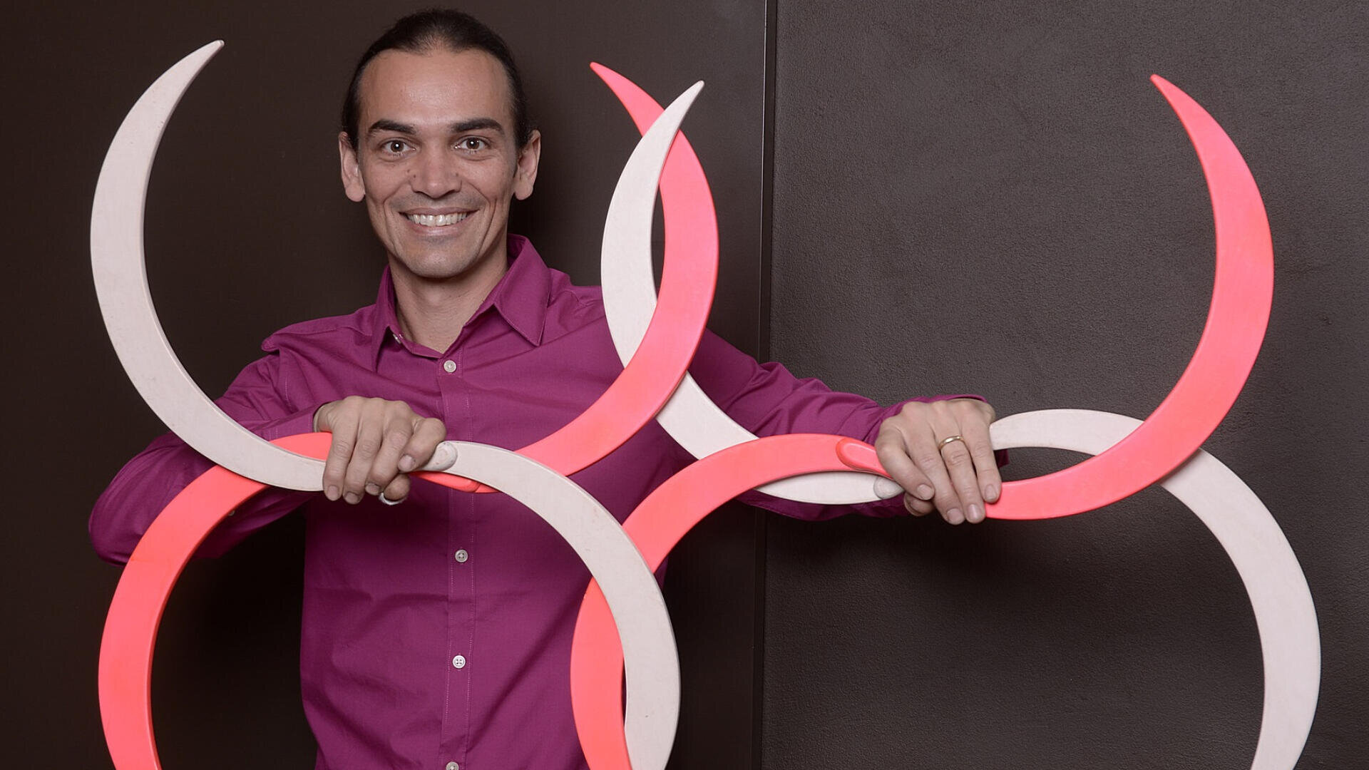 Eine lächelnde Person in einem magentafarbenen Hemd hält vor einem dunklen Hintergrund mehrere ineinandergreifende kreisförmige Objekte, die halb rosa und halb weiß sind.
